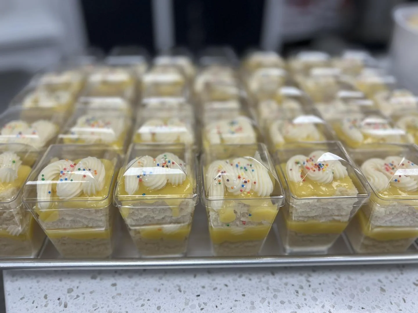 Multiple small plastic cups containing lemon-flavored desserts with whipped cream and colorful sprinkles, arranged on a tray.