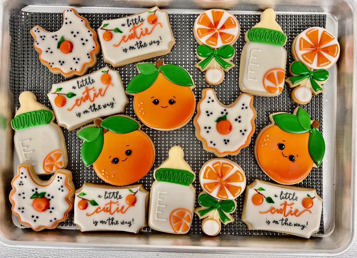 Decorated Christmas cookies on a tray, including oranges with leaves, milk bottles with orange slices, Christmas-themed words, and holly leaves with berries, all with colorful icing.