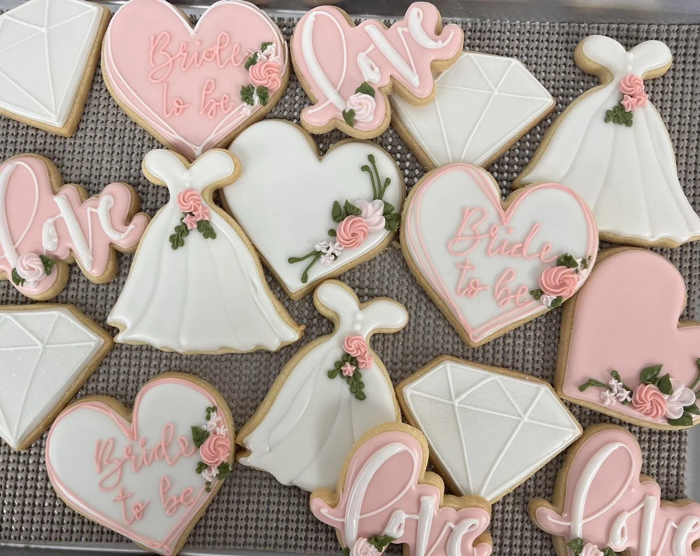 Decorative cookies for a bridal shower, featuring pink hearts with 'Bride to be' written in pink icing, white dresses, and diamond-shaped cookies with white icing, all adorned with pink and green floral accents.