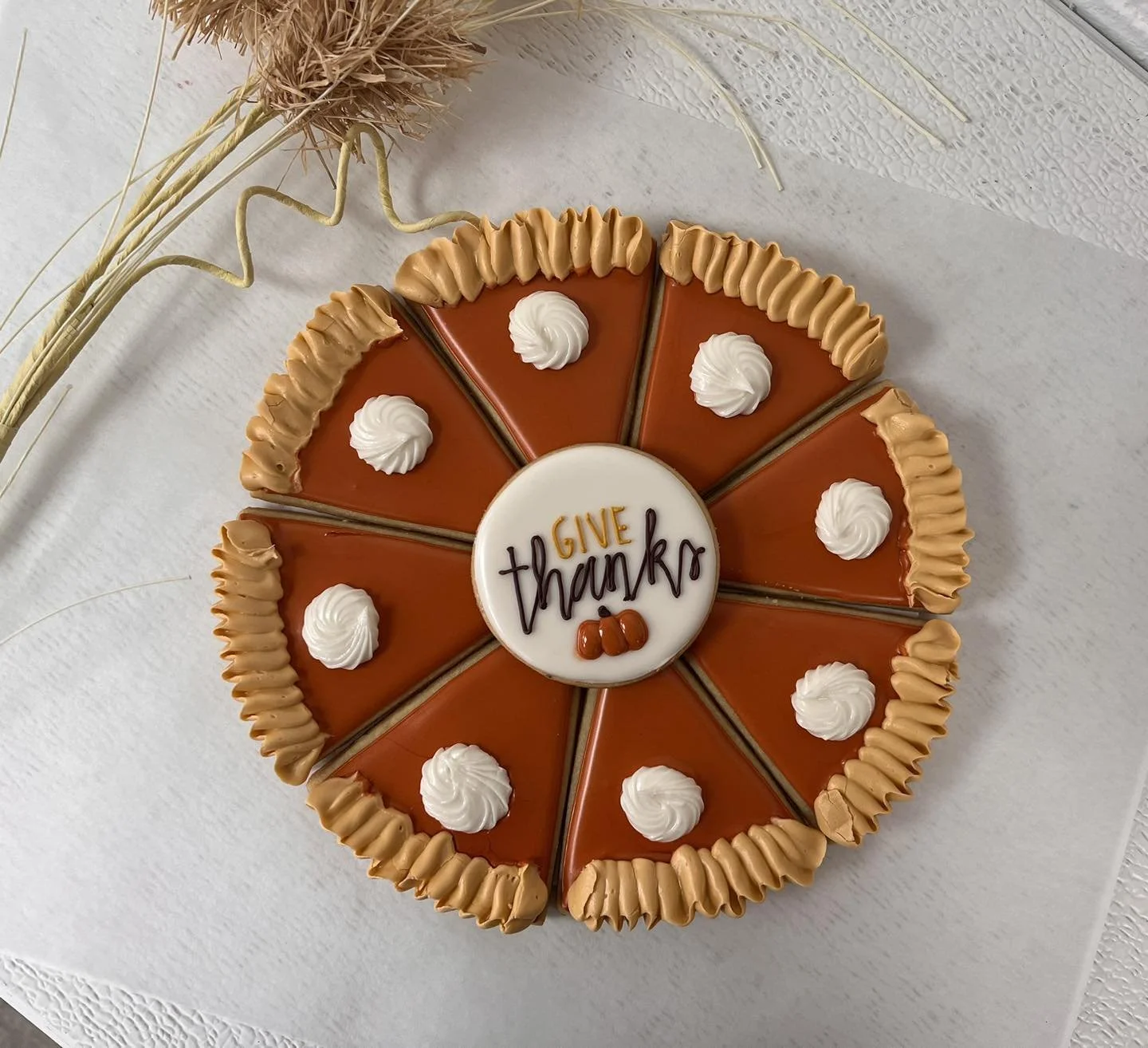 A round caramel-colored pie decorated with white whipped cream dollops and caramel frosting piping around the edge, with a white circular center that says 'GIVE thanks' in black and orange lettering, topped with small caramel candies, on a white surf