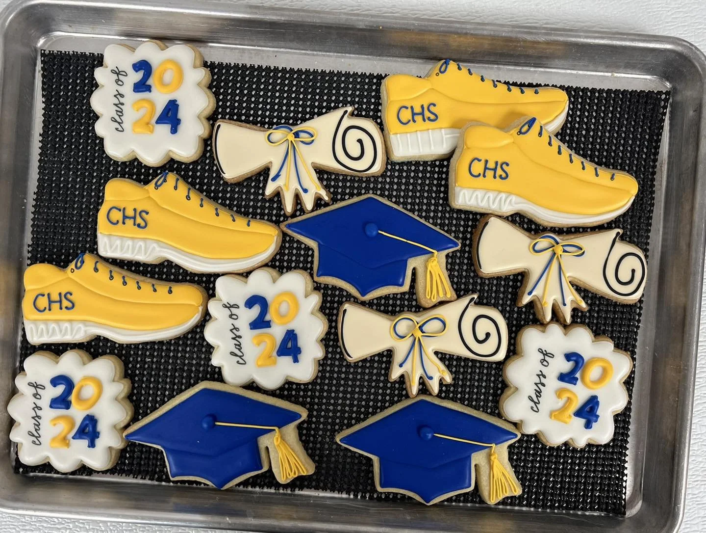 Decorated cookies on a tray celebrating graduation and school achievements. The cookies include yellow sneakers labeled "CHS," white diplomas with blue and yellow "class of 2024," blue graduation caps with yellow tassels, and white scrolls with the y