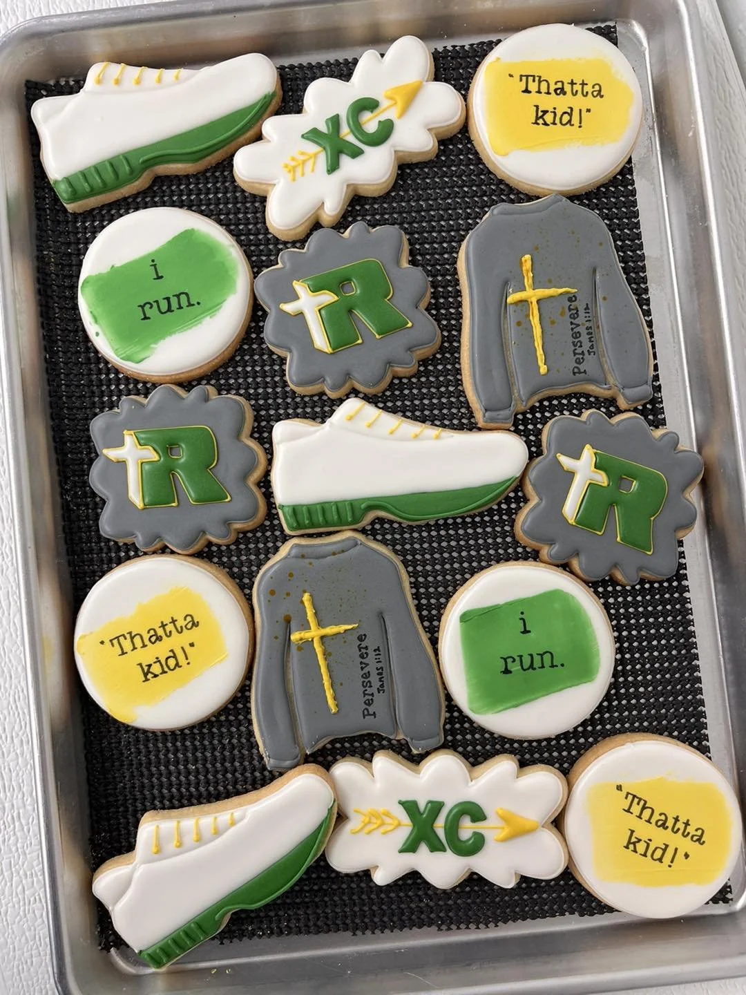Decorated cookies on a black tray with Christian and sports themes. Cookies include a white and green sneaker, gray sweater with a yellow cross and prayer, white and green running shoe, white cookies with green and yellow text saying 'i run.', '