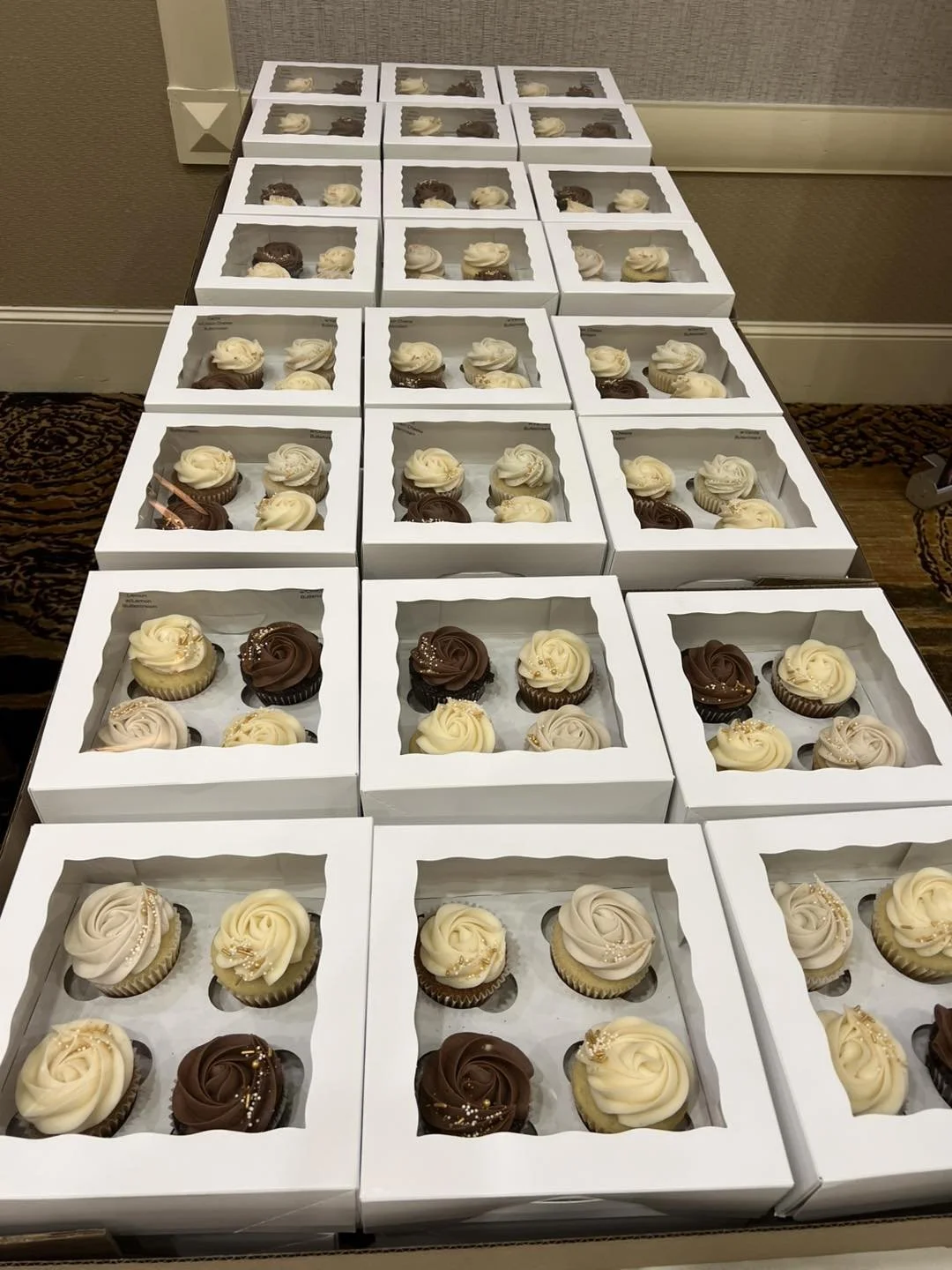 Several boxes of assorted cupcakes arranged on a table, decorated with white, brown, and beige frosting with sprinkles.
