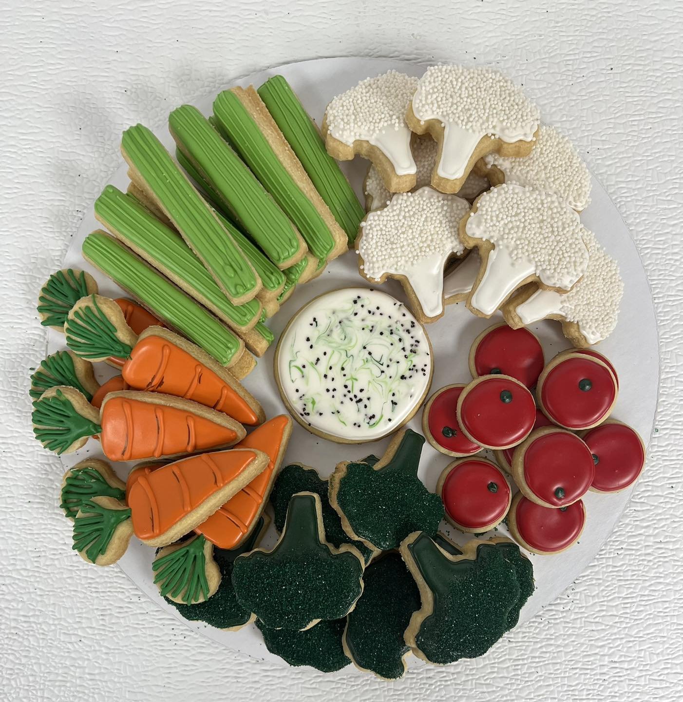 Assorted decorated holiday cookies on a white plate, including carrot-shaped cookies, green and white Christmas trees, white snowflakes, red Christmas ornaments, and a small bowl of frosting with black sprinkles.