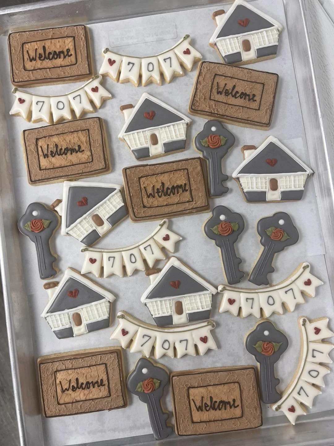 Assorted decorated cookies with a welcome theme, including house-shaped cookies, keys, banners with the numbers 7, 0, 7, and hearts, all arranged on a metal tray.