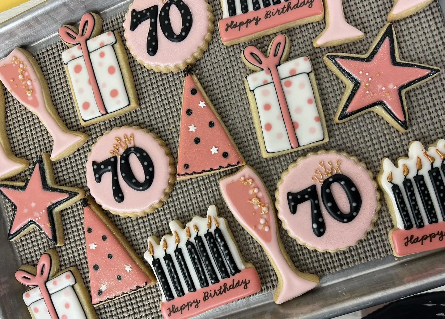 Assorted birthday cookies decorated with pink, black, and white icing featuring birthday-themed shapes like gifts, stars, party hats, cakes, and the number 70