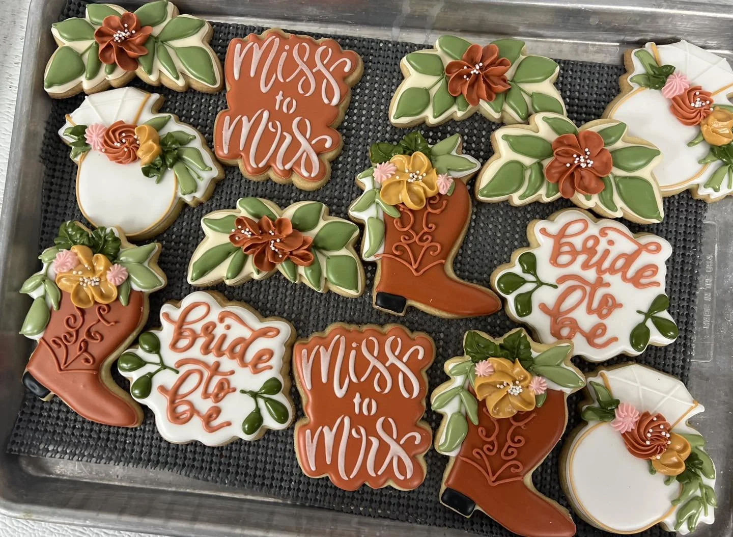 Decorative cookies in various shapes with floral and text designs. Some say 'Miss to Mrs' and 'Bride to be,' others are shaped like flowers, leaves, and a boot, all decorated with icing in colors like white, green, orange, pink, and yellow.