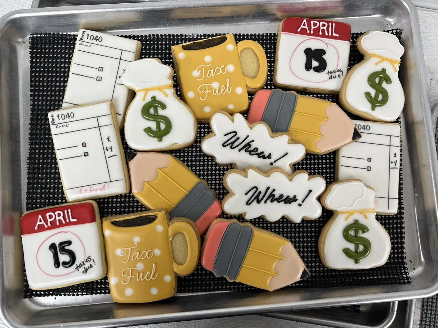 Assorted decorated cookies on a metal tray, featuring images of money bags, coffee mugs, pencils, invoices, and messages like 'Whew!' and 'Tax Fuel'.