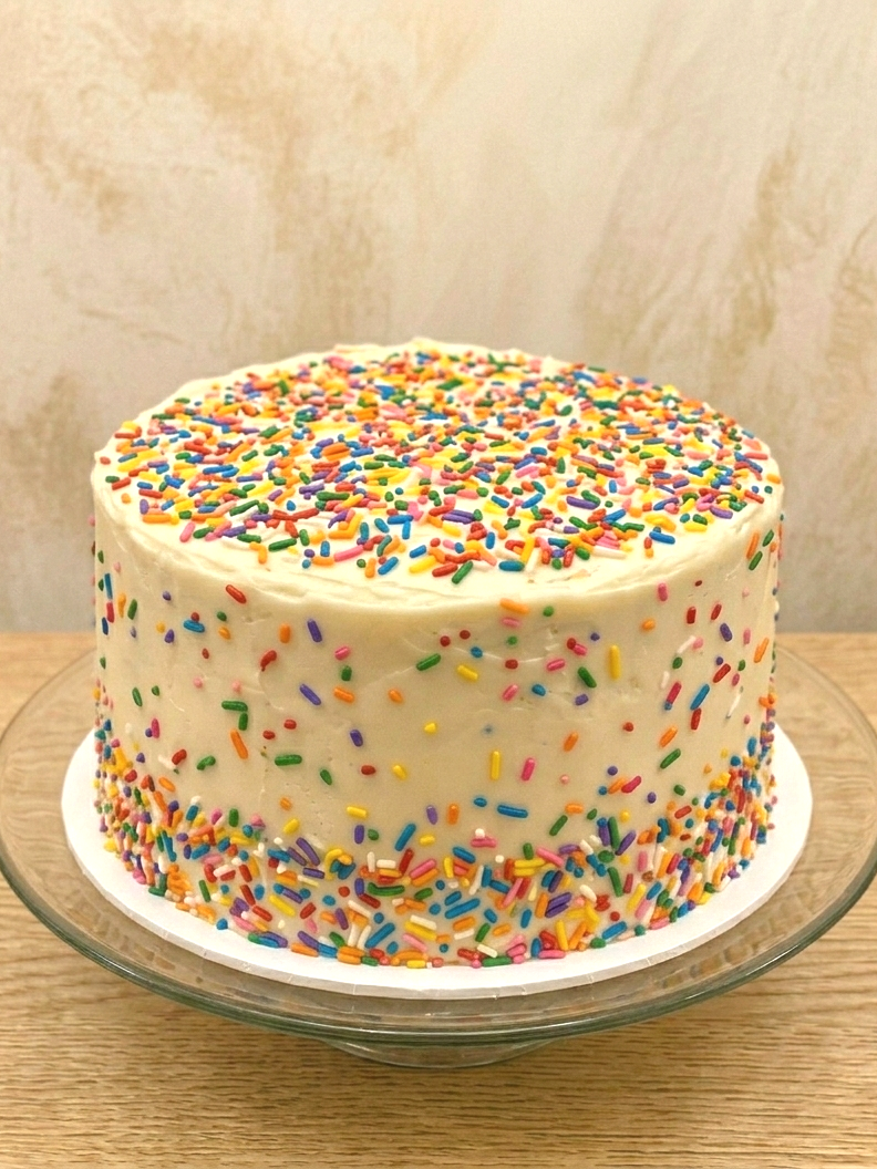 Celebration cake with white frosting and colorful rainbow sprinkles on top and around the sides, placed on a glass cake stand on a wooden surface.