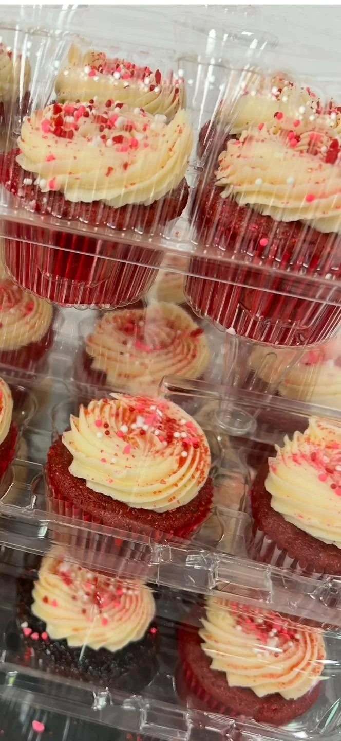 Red velvet cupcakes with cream cheese frosting and pink and red sprinkles in clear plastic containers.