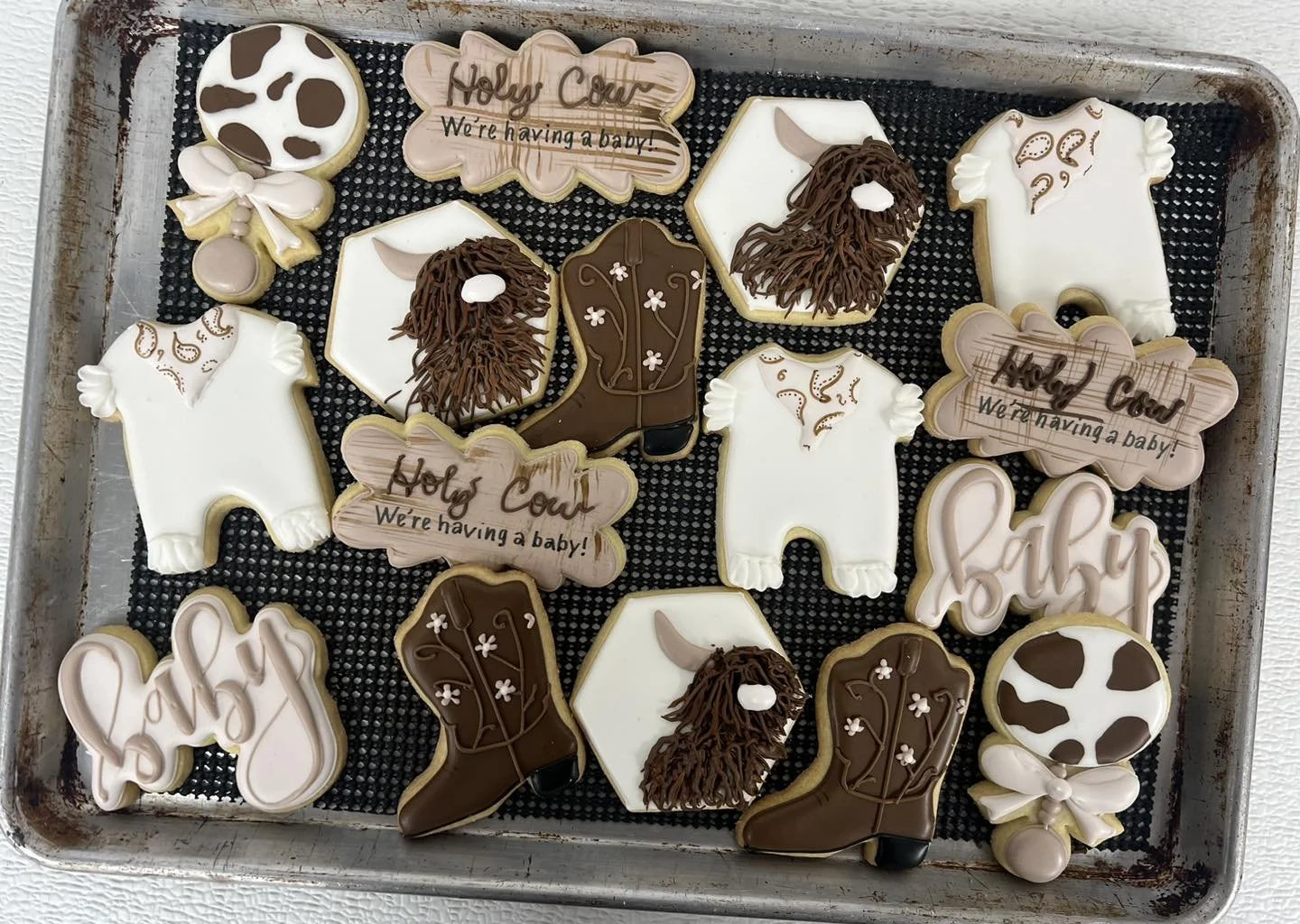 Assorted decorated cookies with baby shower theme, including ones shaped like boots, animals, hearts, and clouds, some with messages like 'Holy Cow! We're having a baby!' and decorated in white, brown, and beige icing.
