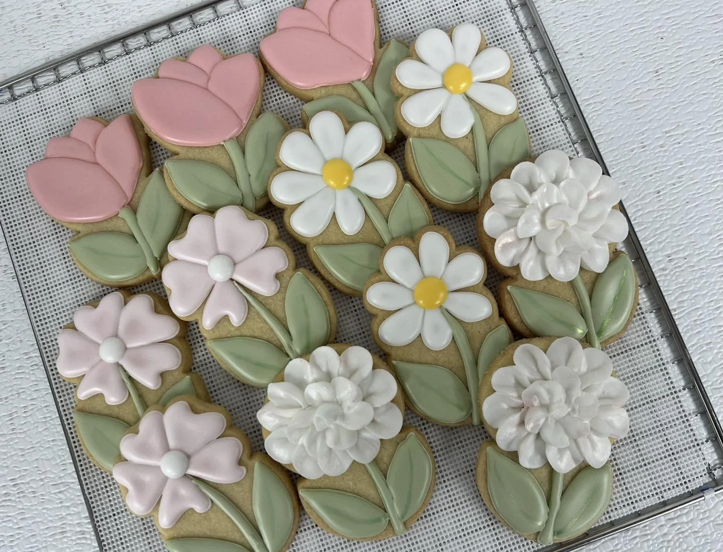Decorative cookies shaped and decorated as pink tulips, white daisies with yellow centers, pink flowers with white centers, and white carnations with green leaves, arranged on a cooling rack.