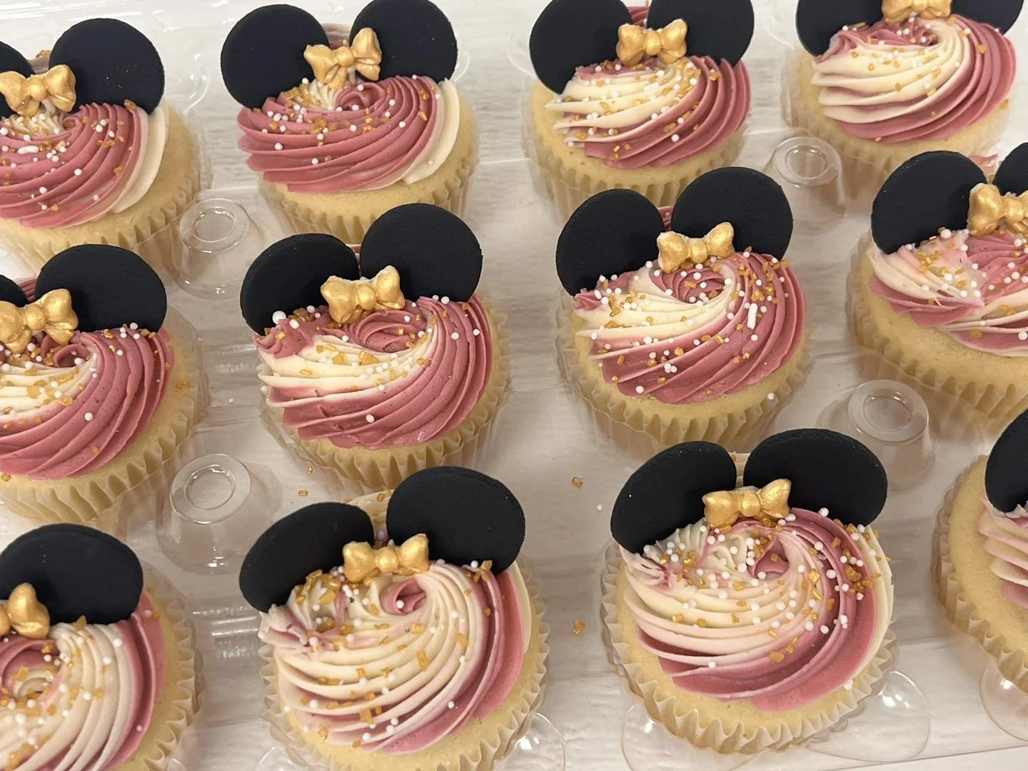 Nine decorated cupcakes with pink and white swirl frosting, black cookie ears, and a small gold bow decoration arranged on a clear plastic tray.