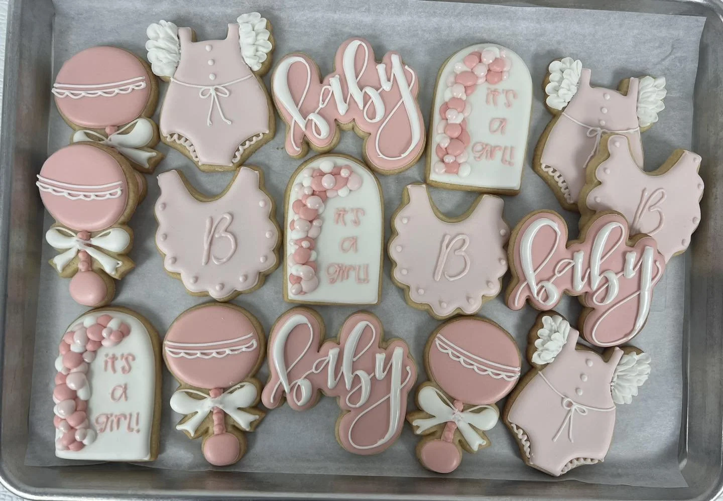 Assorted baby shower cookies decorated with pink, white, and light purple icing, including ones shaped like baby onesies, baby bottles, balloons, and plaques with messages such as 'It's a girl!' and 'Baby'.