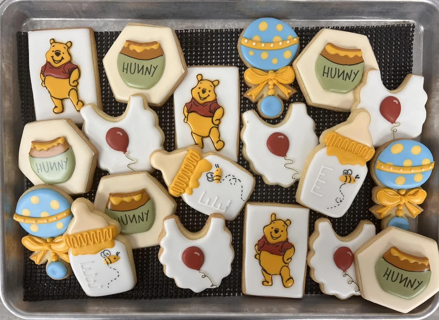Assorted decorated cookies with Winnie the Pooh, honey jars, balloons, and bee designs on a baking tray.