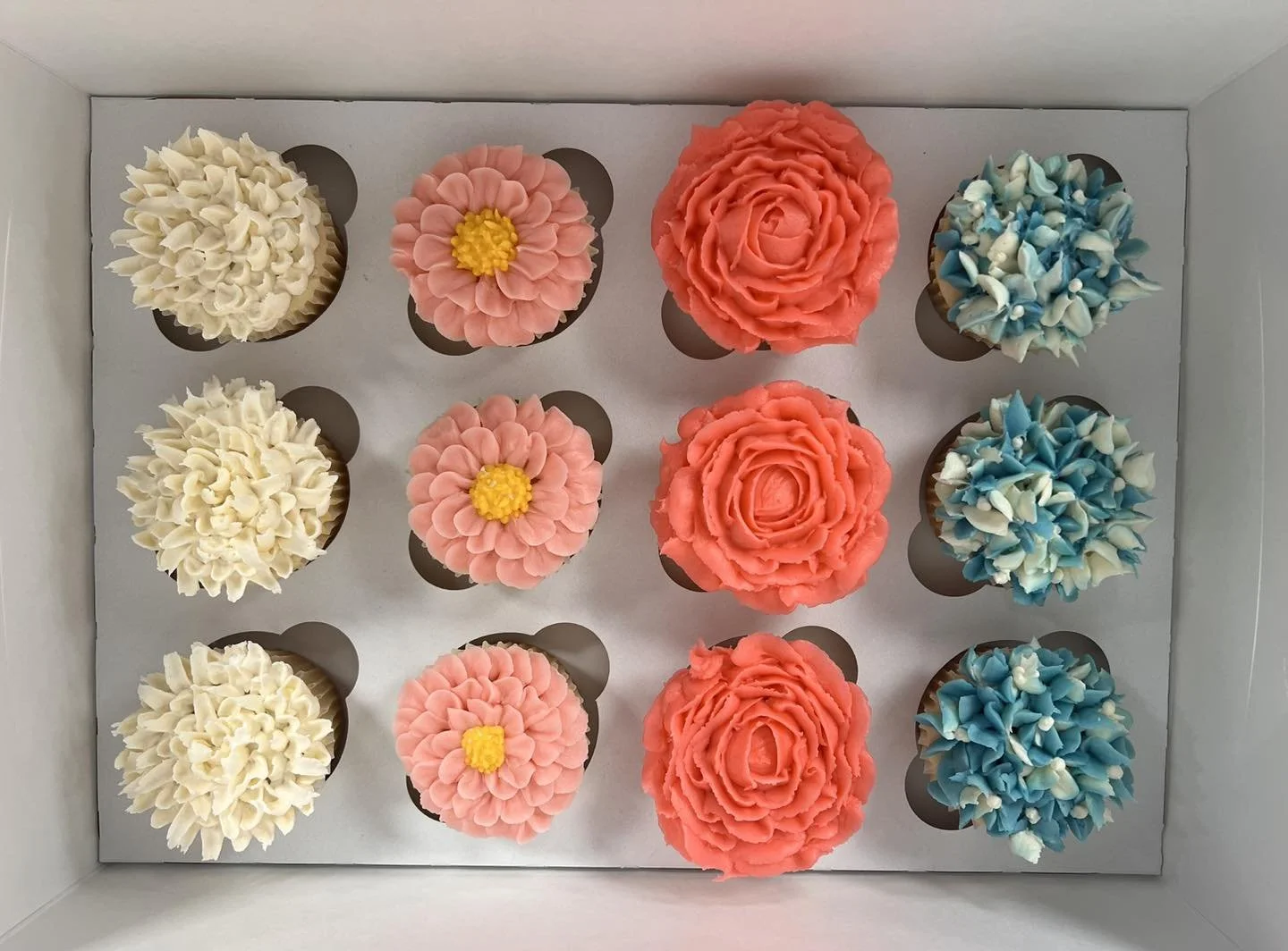 A box of twelve cupcakes decorated with different floral-themed frosting in white, pink, coral, and blue colors.