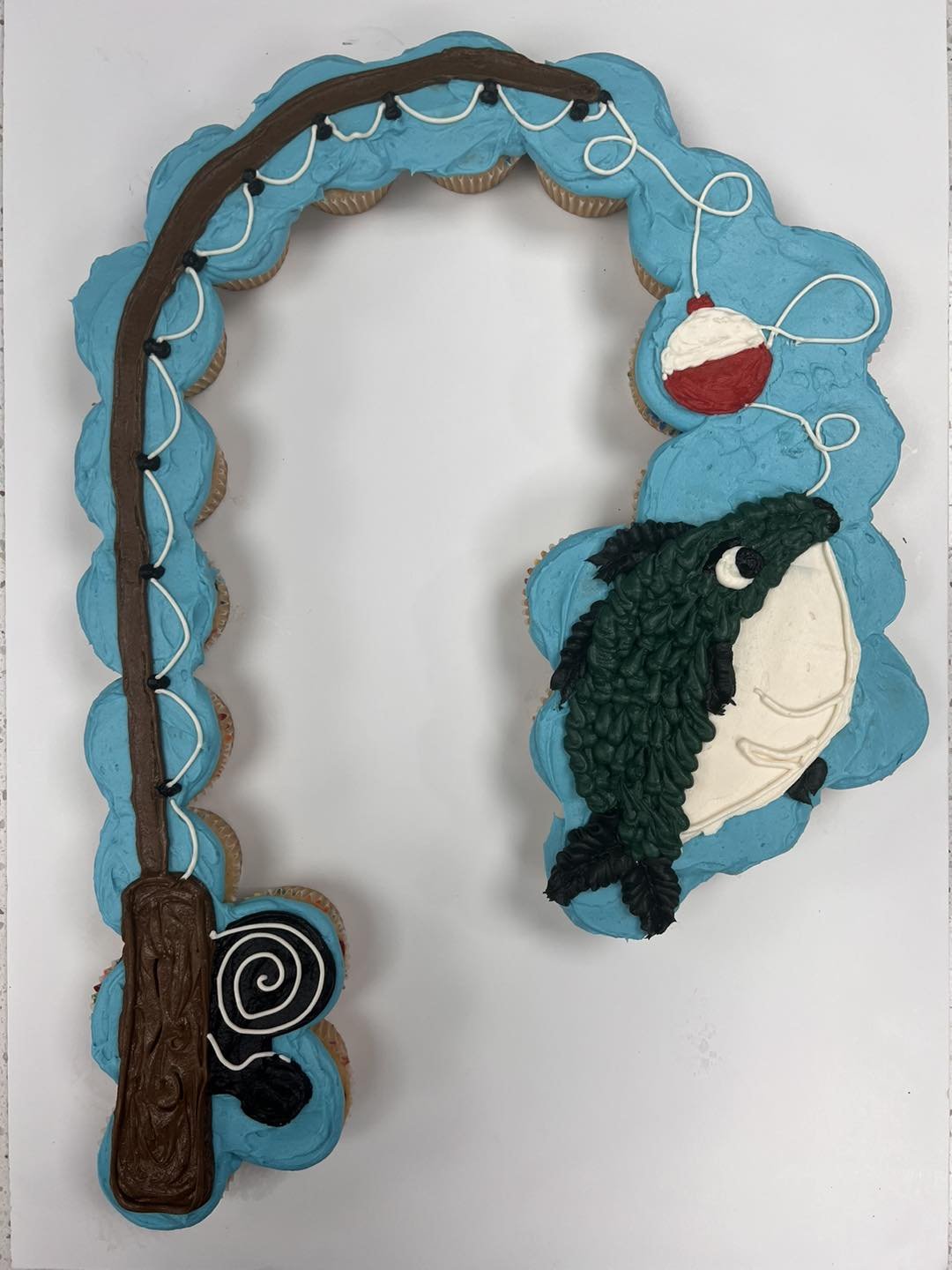 Cake decorated with blue icing, a fishing rod made of brown and black icing, a red and white bobber, and a black fish with a white eye and cream-colored belly, designed to resemble a fishing scene.