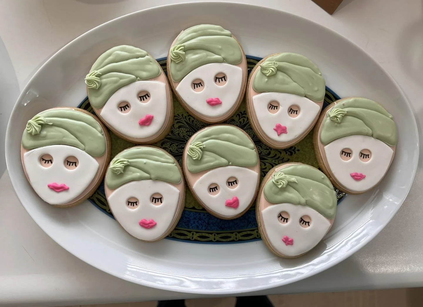 A white oval platter holding eight cookies decorated as stylized female faces with green hair, white skin, closed eyes, and pink lips.