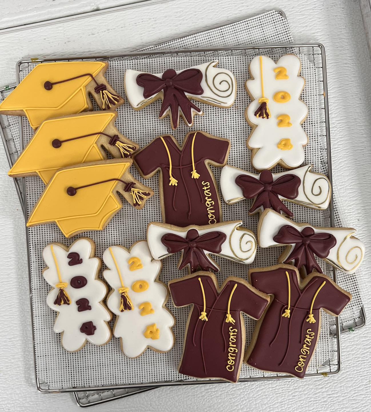 Assorted graduation-themed cookies on a cooling rack, including diploma scrolls, graduation caps, and mortarboard cookies with yellow and maroon decorations.