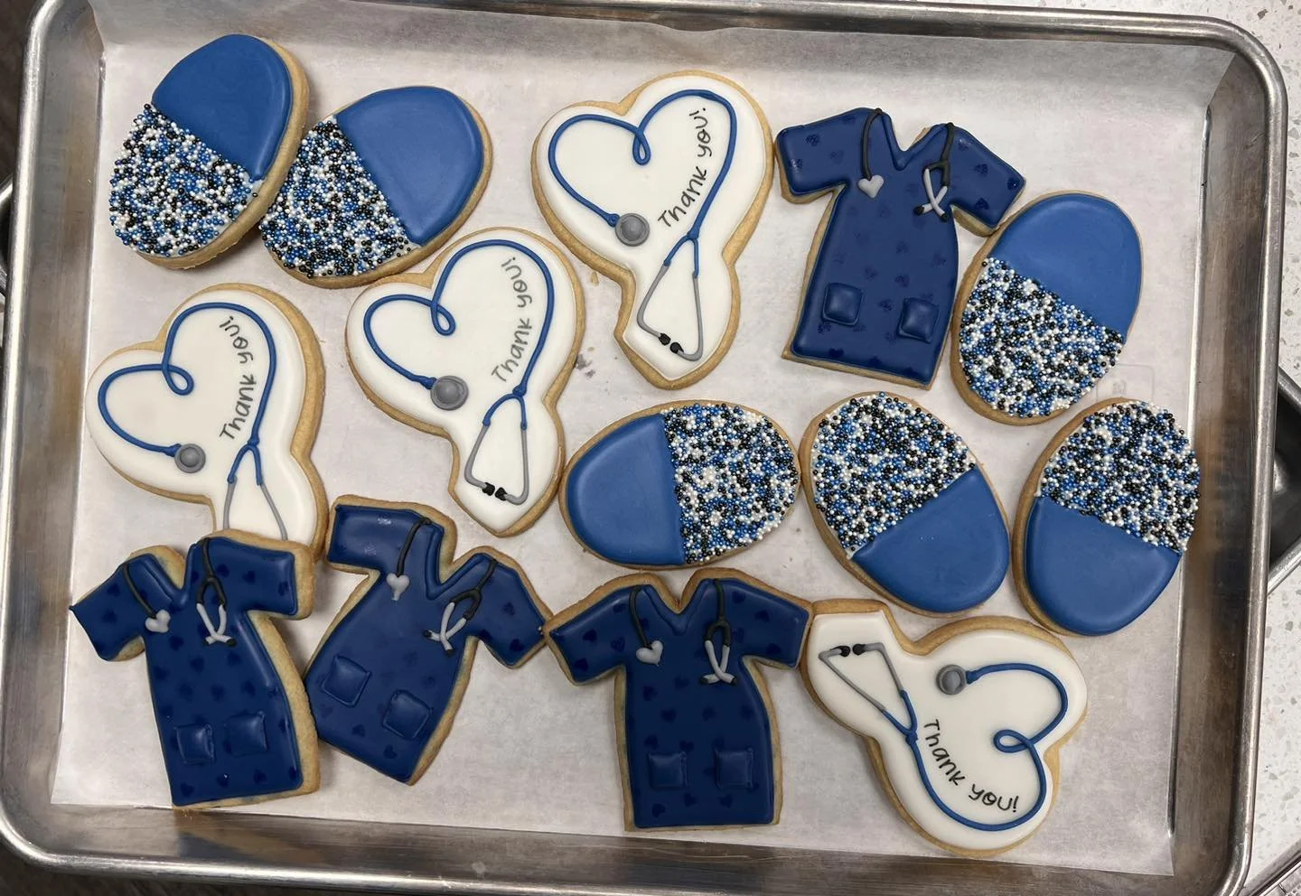 Decorated cookies with medical themes, including hearts with stethoscope designs, hospital gowns, and pills, in blue, white, and silver colors on a baking sheet.