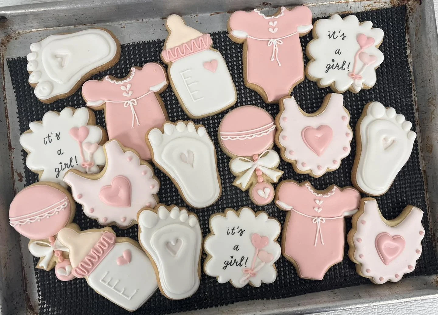 Decorated cookies celebrating the arrival of a girl, shaped as pink baby onesies, milk bottles, baby rattles, bibs, pacifiers, and a balloon, with inscriptions like "it's a girl!"