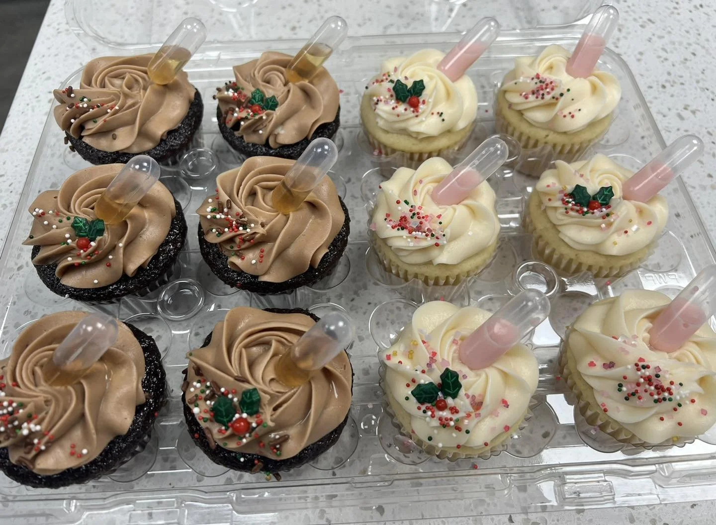 Cupcakes decorated with holiday sprinkles, holly leaves, red berries, and topped with pink or brown liquid-filled syringes in clear plastic stands.