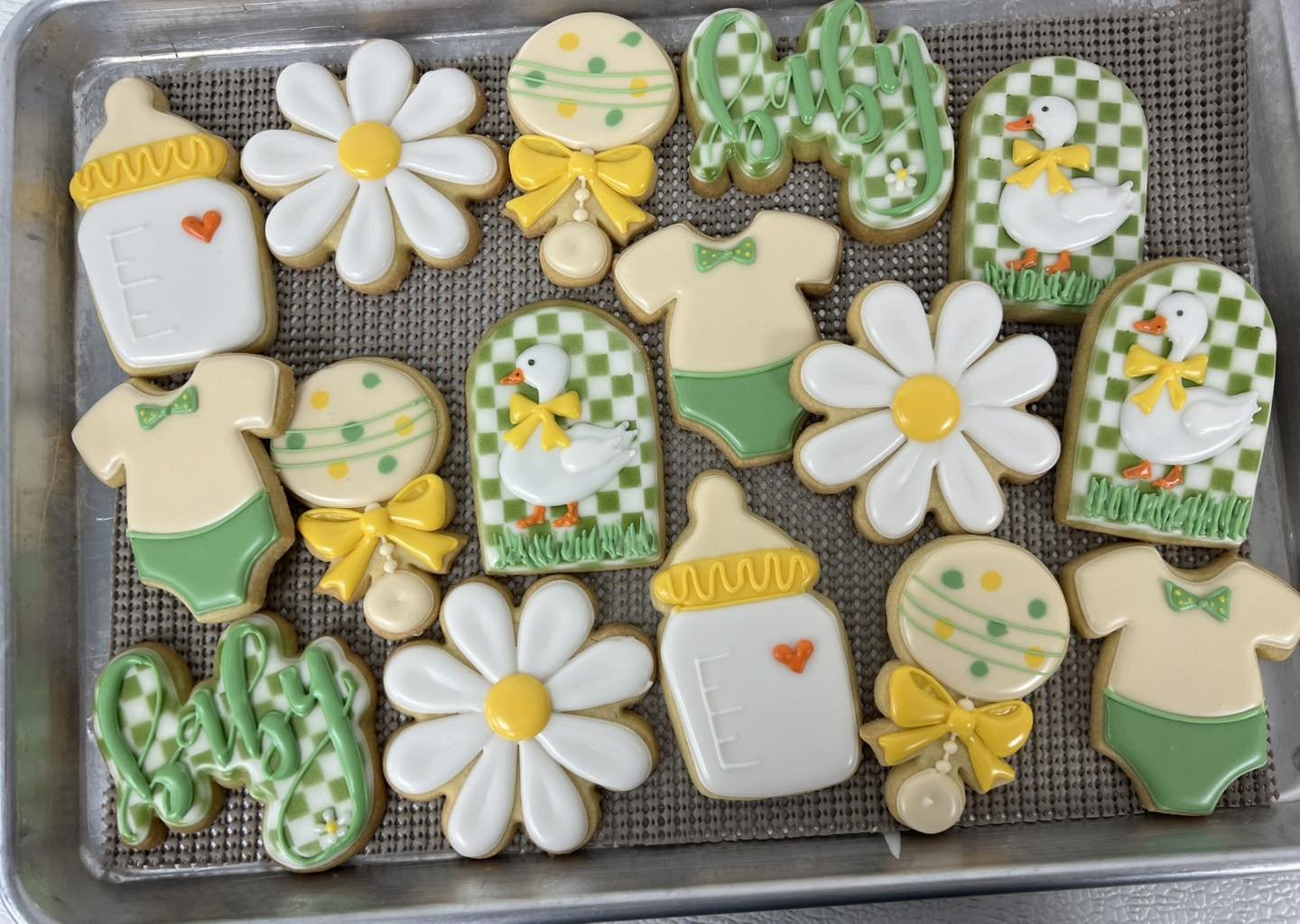 Decorated sugar cookies with baby shower themes, including bottles, onesies, flowers, ducks, and balloons in pastel green, yellow, and white colors.