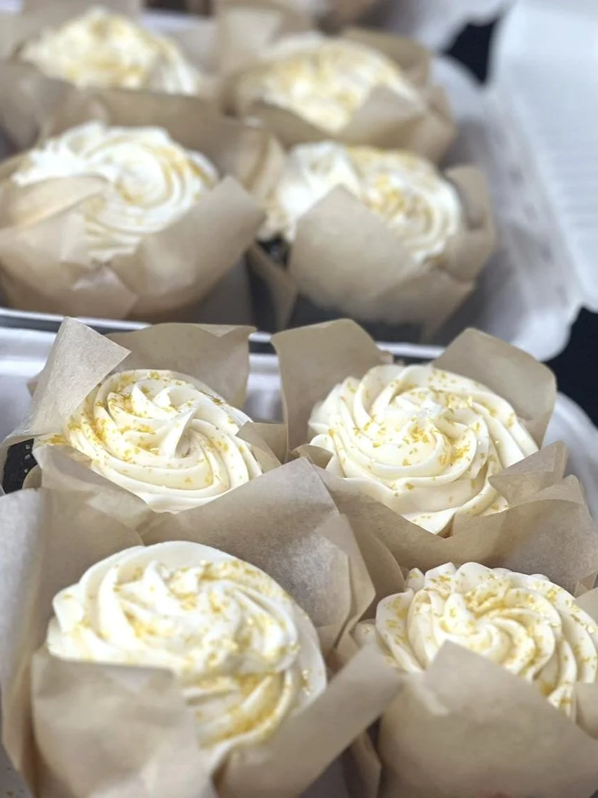 A tray of cupcakes decorated with white frosting and yellow sprinkles, each in parchment paper cupcake liners.