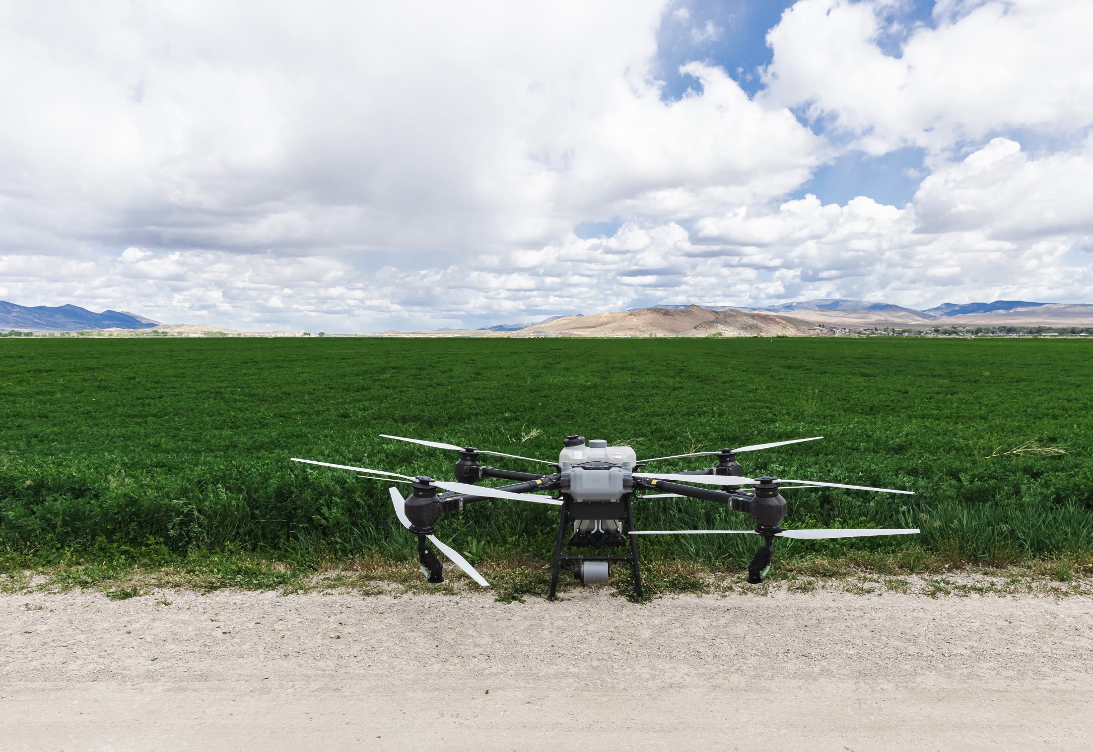 An agricultural drone stationed on a dirt path in front of a lush green field, with a mountain range and cloudy sky in the background.