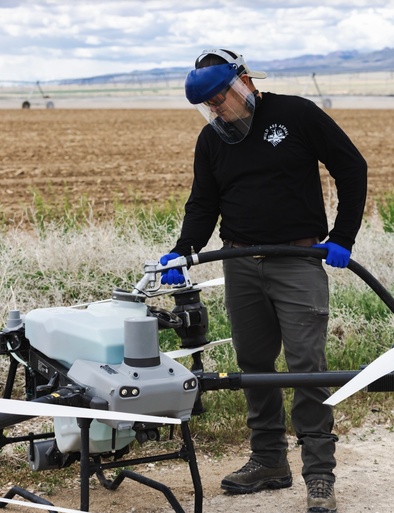 A person wearing safety goggles, a face shield, and gloves operates a drone in an open field with farmland and mountains in the background.