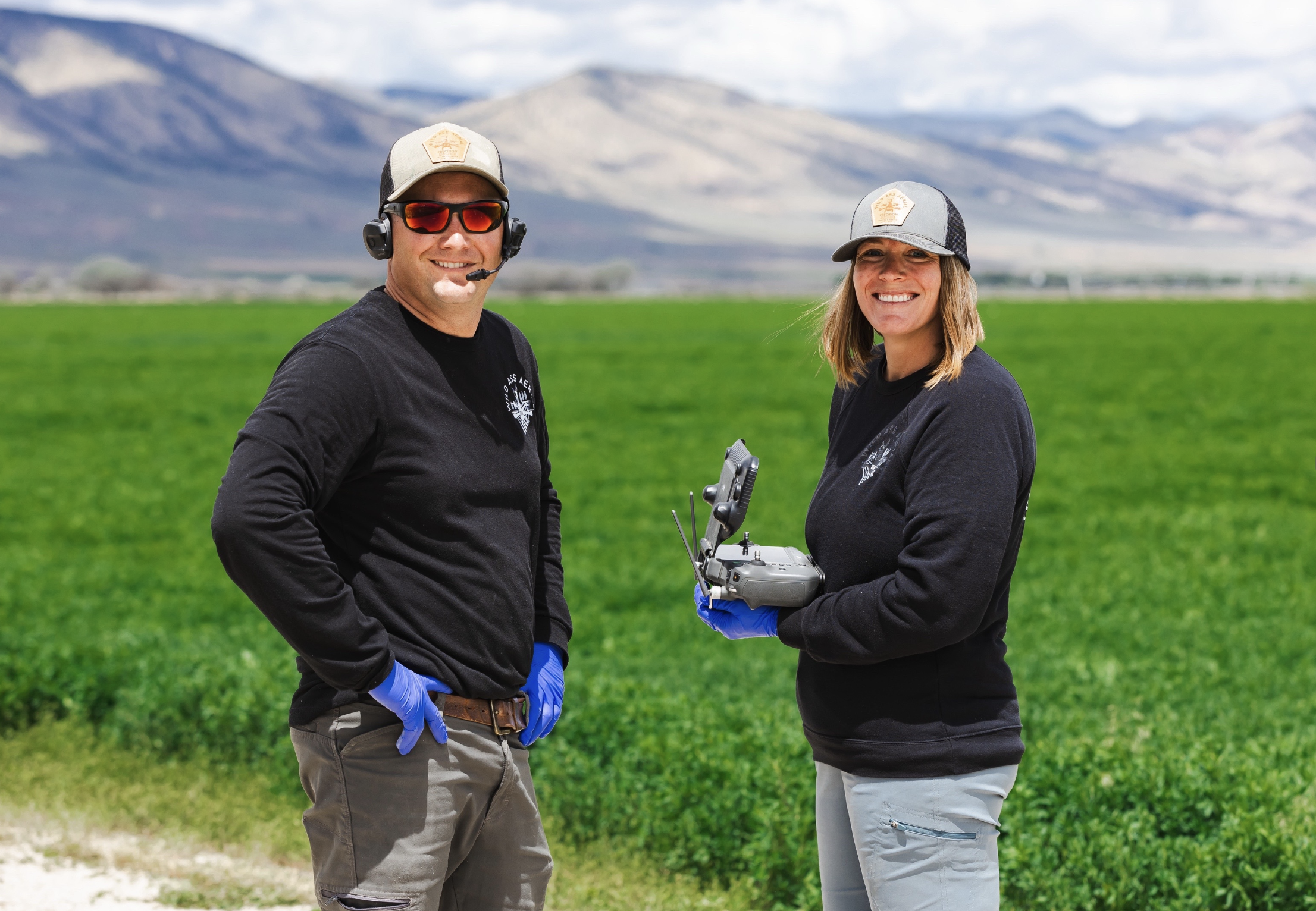 Two people wearing black shirts, gloves, and hats standing in a green field with mountains in the background. One is holding a drone remote controller. They are smiling at the camera.