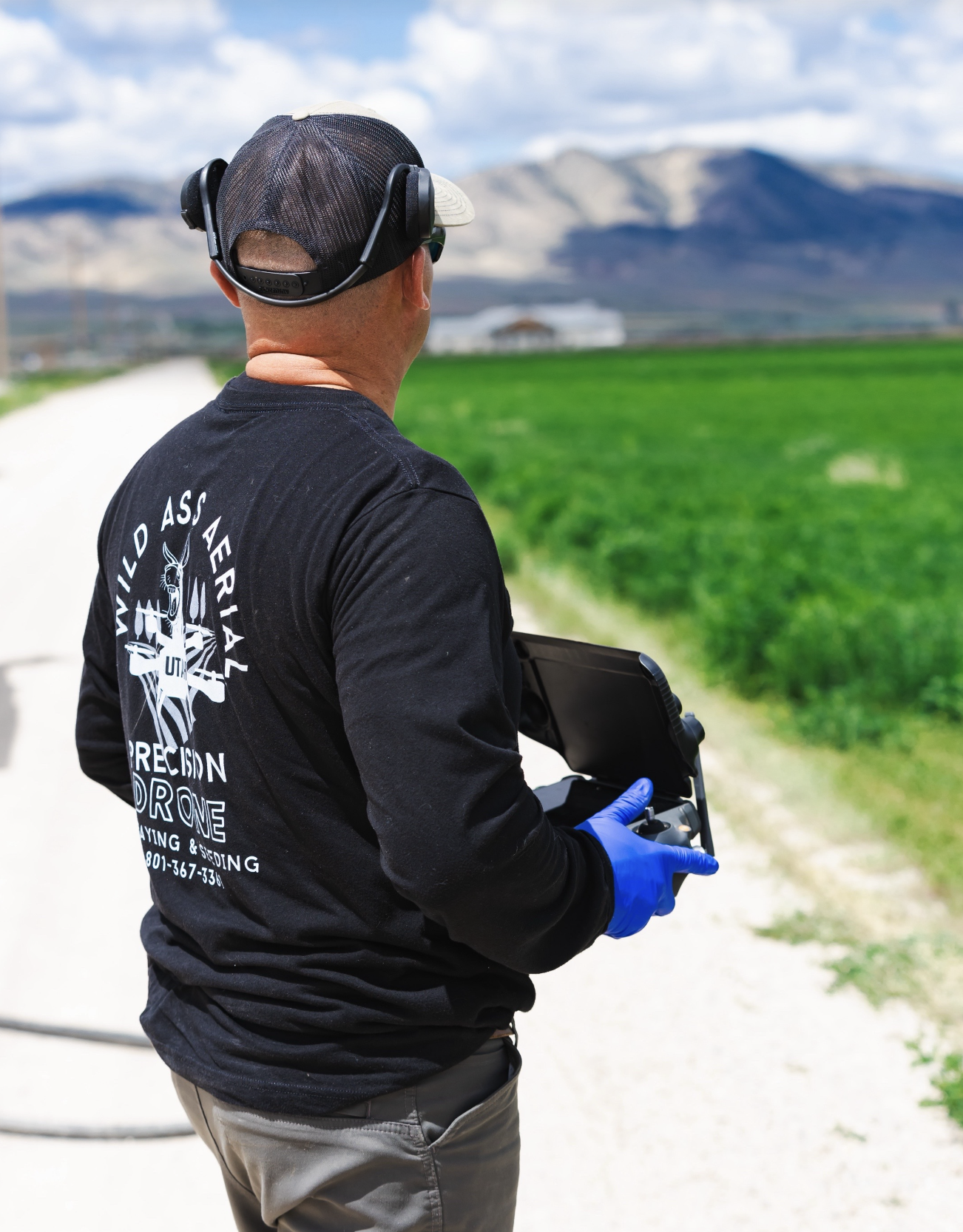 Man flying a drone in a rural area with green fields and mountains in the background, wearing a black shirt, a cap, headphones, and gloves, holding a remote control.