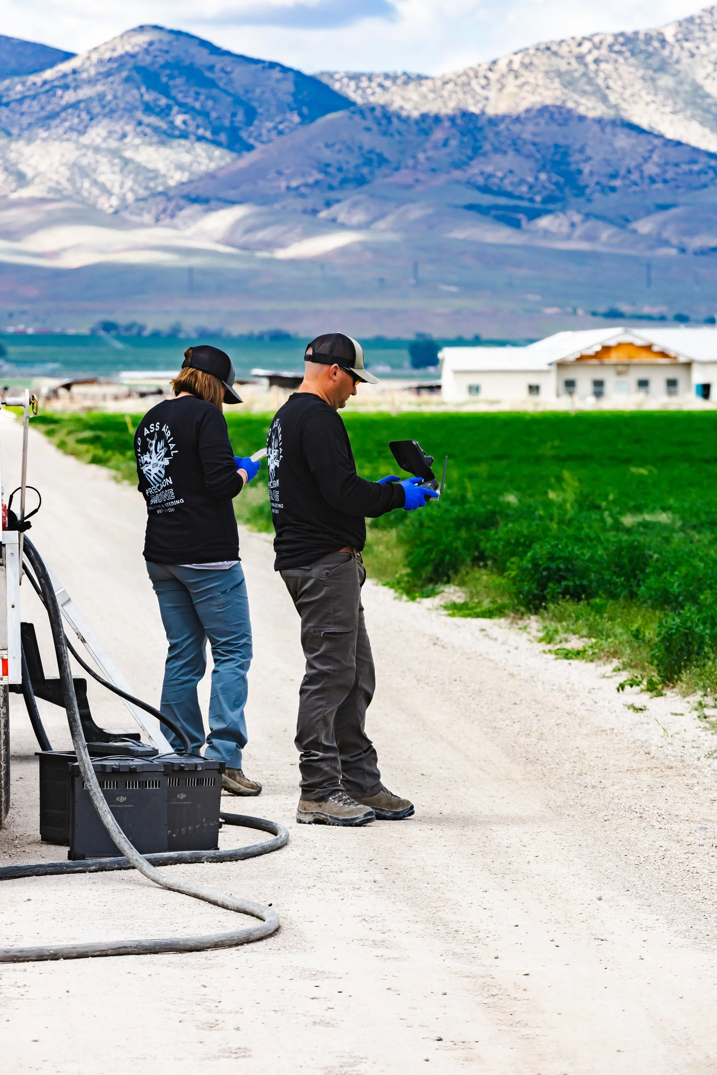 Two people in black jackets and hats standing on a dirt road on a farm, operating drone equipment with a backdrop of mountains and green fields.