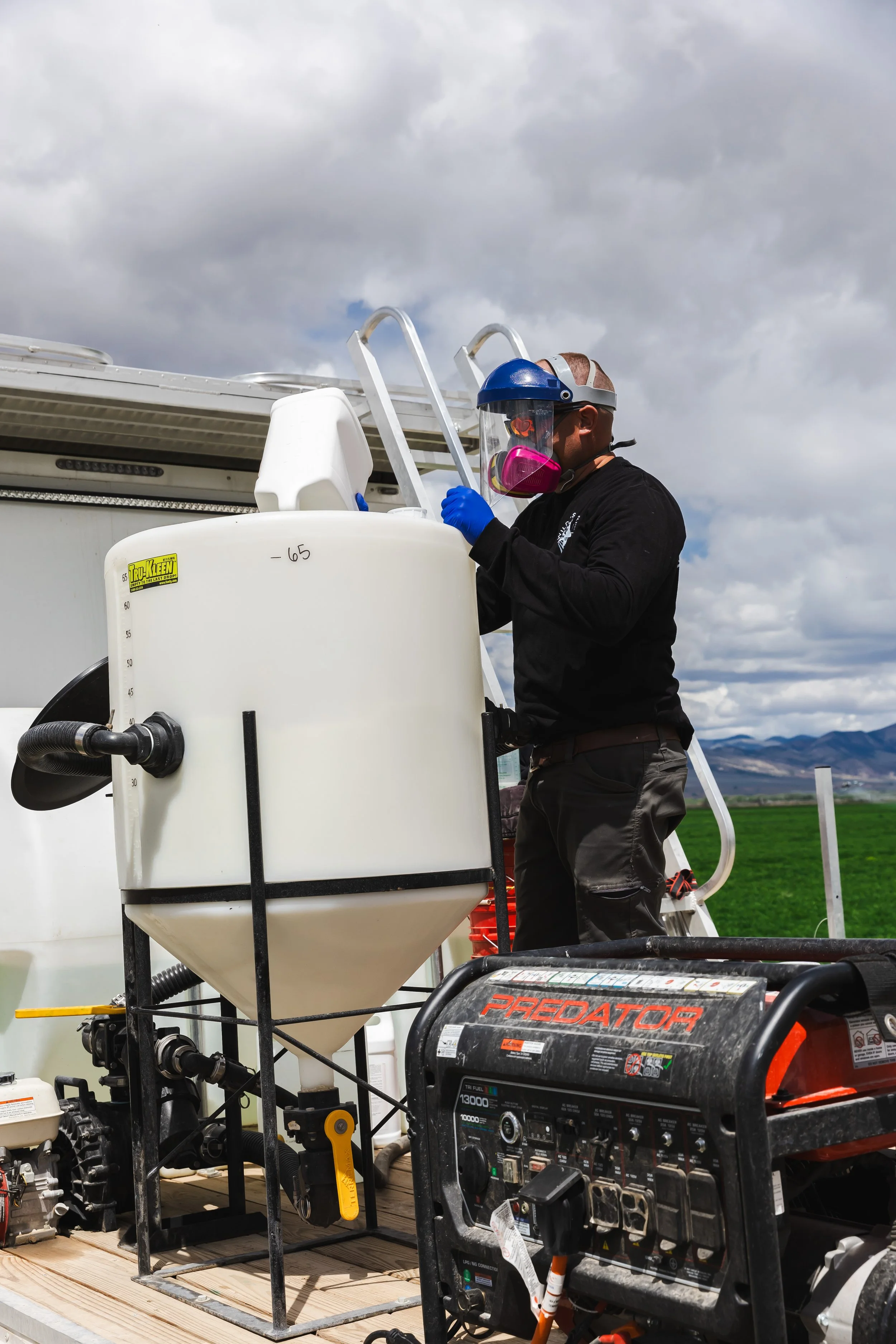 A worker wearing a face shield, respirator mask, gloves, and black shirt working on equipment on a farm or field with mountains and cloudy sky in the background.