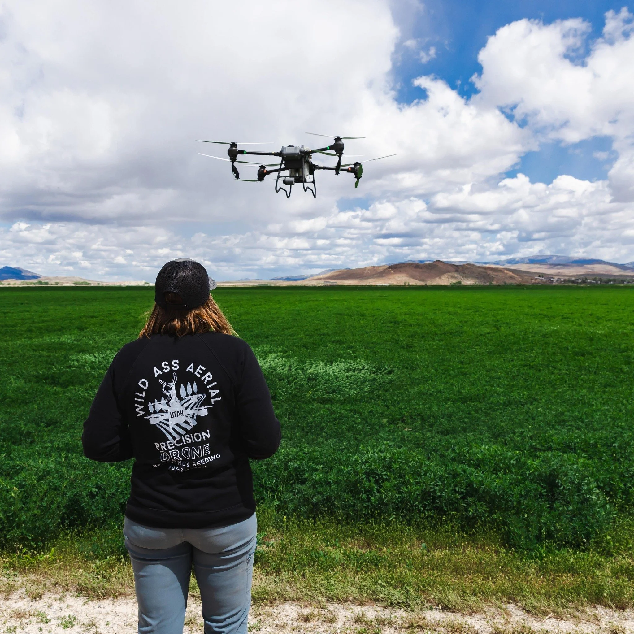 A person in a black jacket with a white logo on the back watching a drone fly over green fields under a partly cloudy sky.