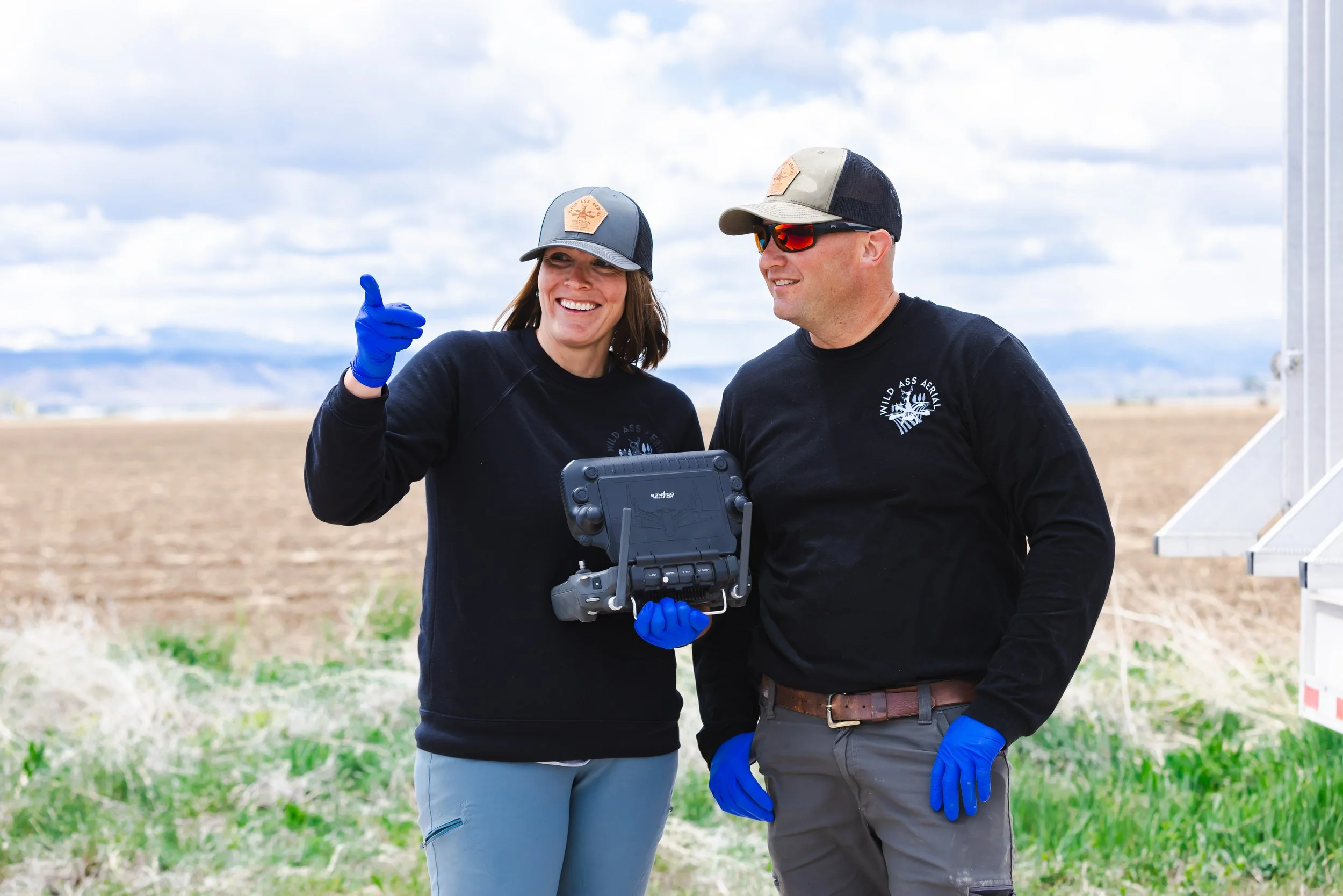 Two people wearing black sweatshirts and hats with sunglasses and blue gloves are standing in a field, smiling and talking to each other, with a tablet in hand. One person is pointing, and the background shows a cloudy sky and open land.