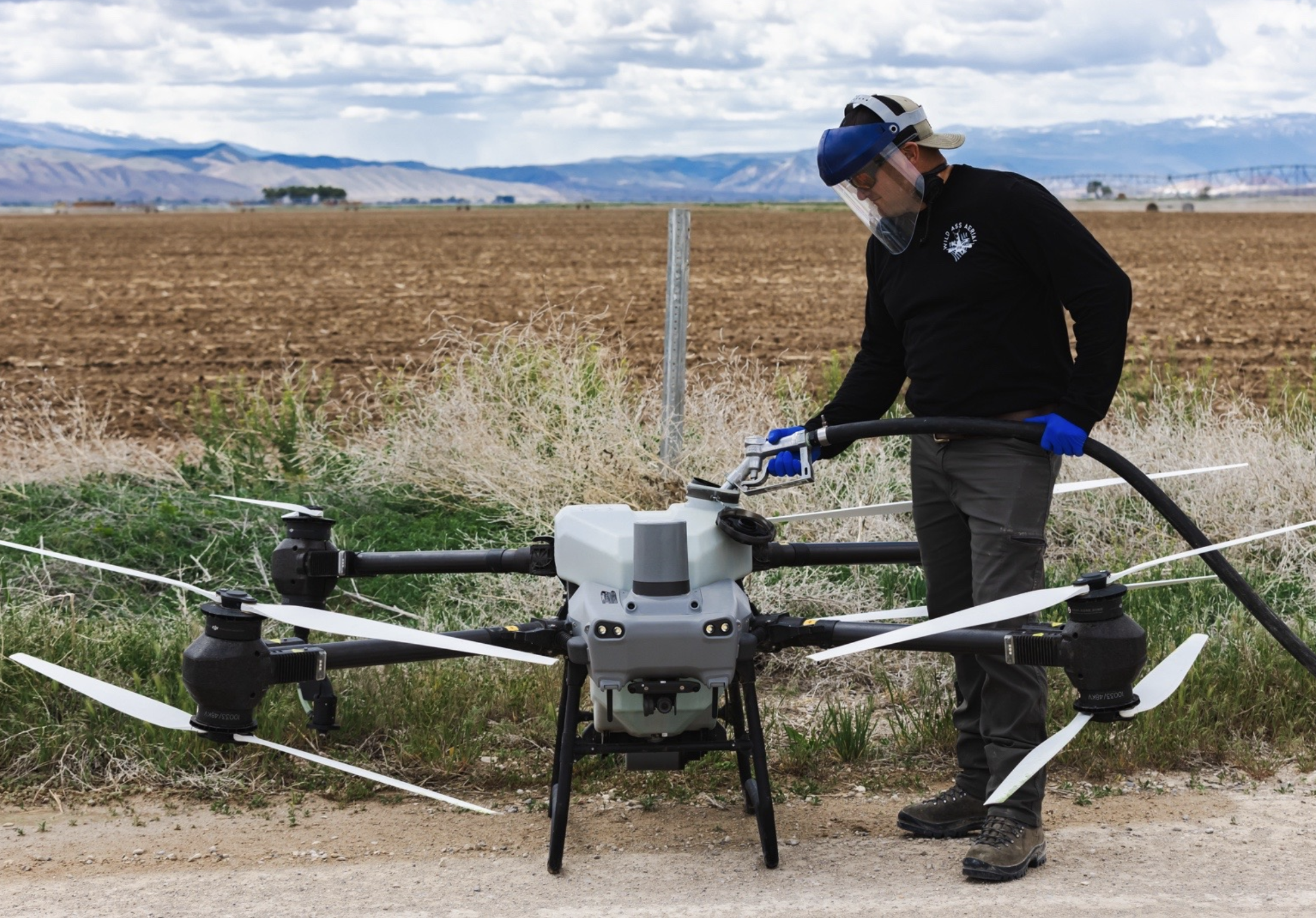 A person in protective gear operating an agricultural drone in a field with mountains in the background.