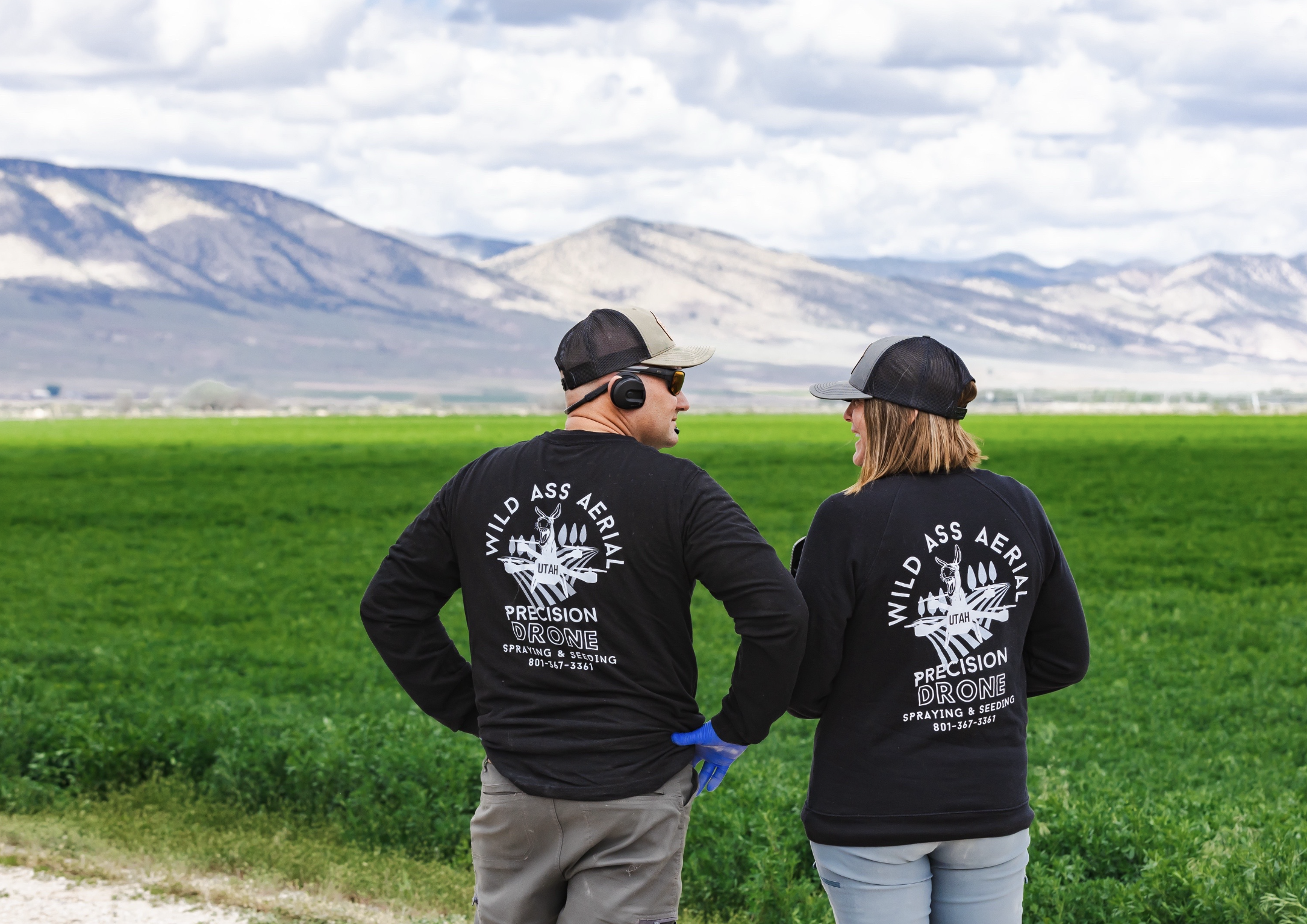 Two people in black sweatshirts with 'Wild Ass Aerial Precision Drone Spraying & Seeding' and a graphic of a drone, standing in a lush green field with mountains in the background, talking to each other.