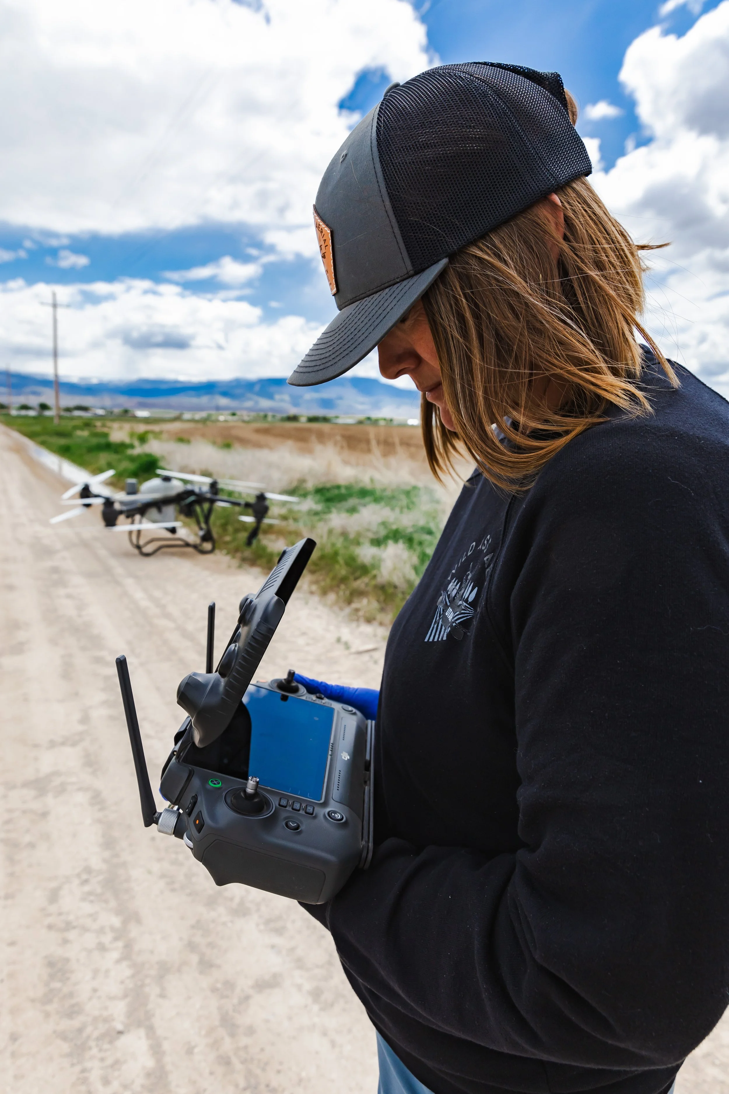 A woman in a black jacket and cap holding a drone remote controller, with a drone on the ground nearby in a rural area with a dirt road and cloudy sky.