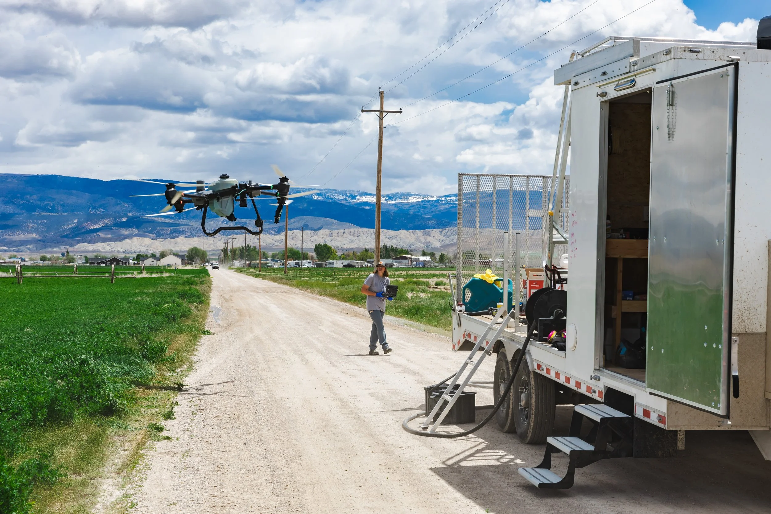 A worker standing on a dirt road with a drone flying nearby. There is a mobile work truck with equipment and a ladder, with mountains and a cloudy sky in the background.