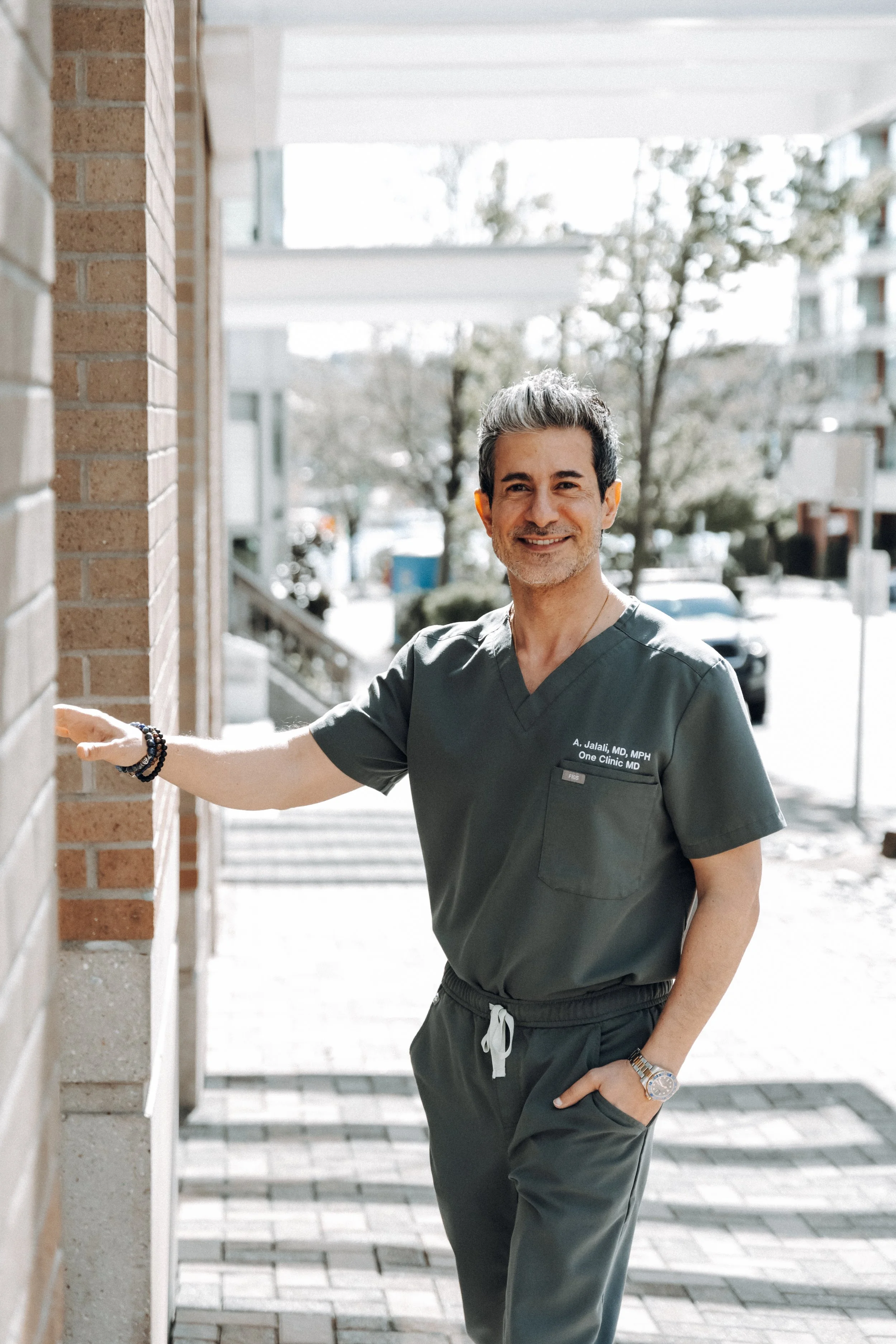 A smiling male healthcare professional in gray scrubs standing outside near a brick wall with a hand resting on the wall.