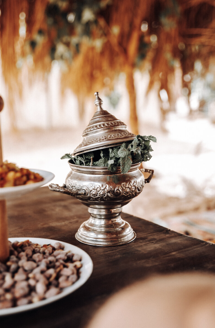 A traditional Middle Eastern or Indian silver serving dish with a domed lid, containing fresh mint leaves, on a wooden table. Part of a plate with food is visible in the foreground, on a blurred outdoor setting with trees in the background.