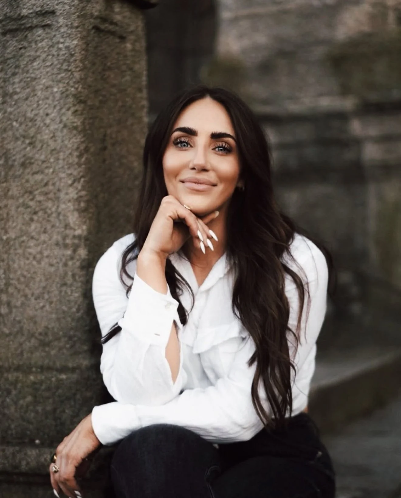 A woman sitting against a stone wall wearing a white shirt, with long dark hair, smiling softly and resting her chin on her hand.