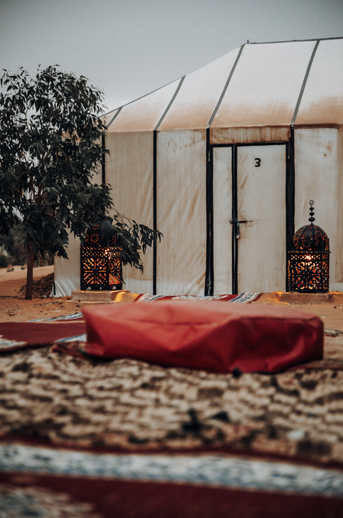 Inside a desert tent with a small tree, two lit lanterns, colorful carpets, and a red pillow or cushion.