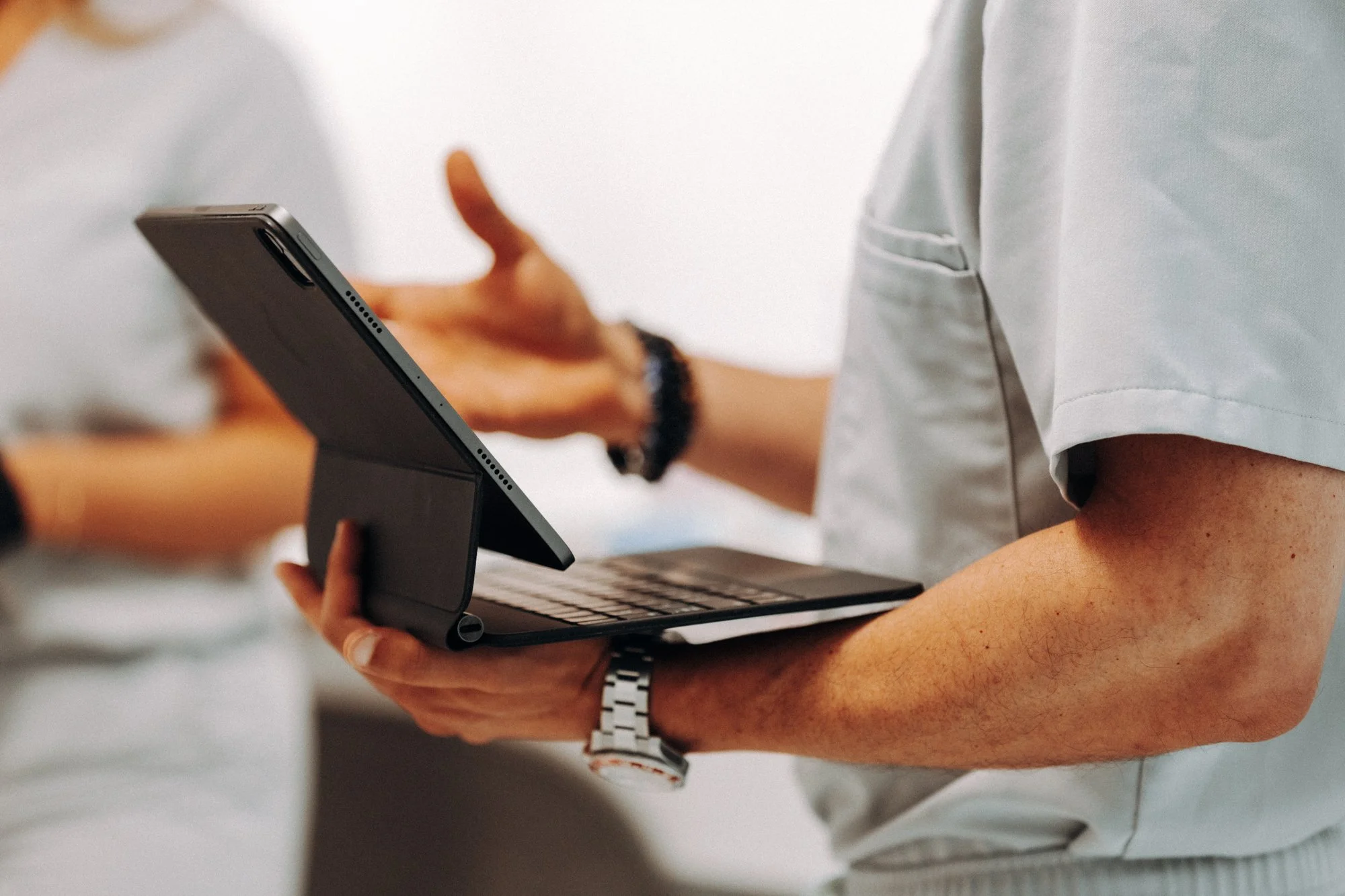 A person in a white shirt holding a tablet computer with a keyboard case, gesturing with their other hand in a discussion.