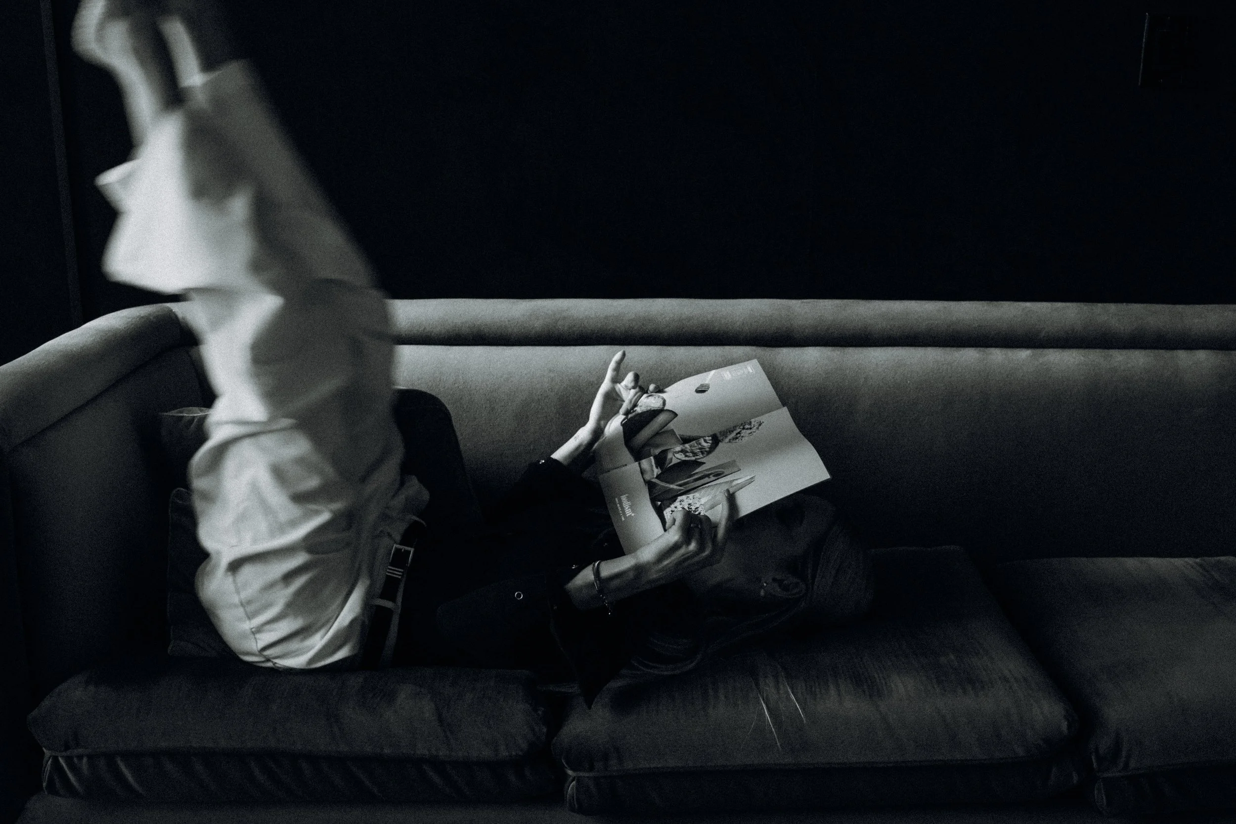 A person lying on a dark sofa, reading a magazine in a dimly lit room, with their legs resting on the armrest and their face obscured by the magazine.