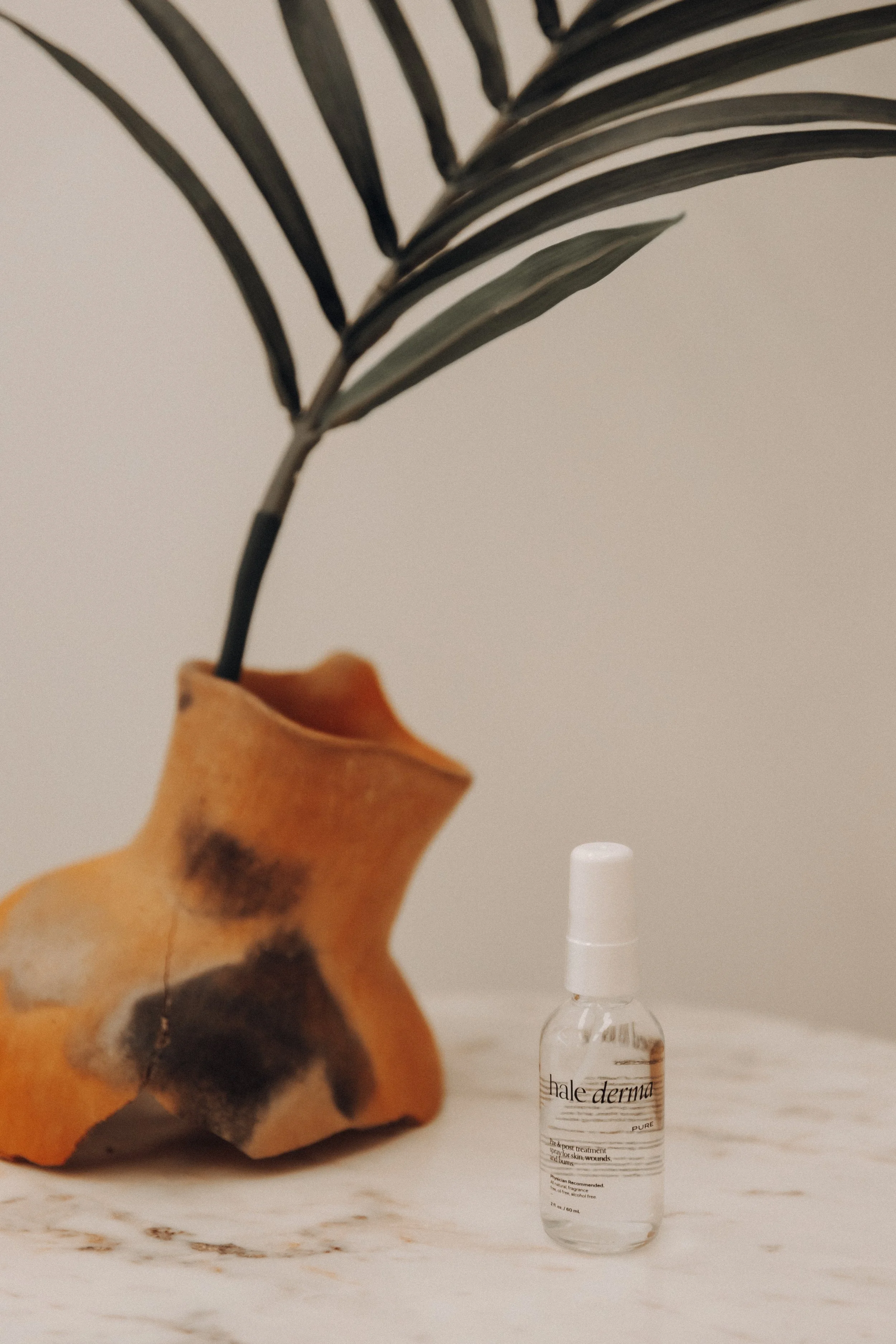 A small spray bottle labeled 'halee derma' placed on a white surface next to a decorative ceramic horse head and a green leafy plant in the background.