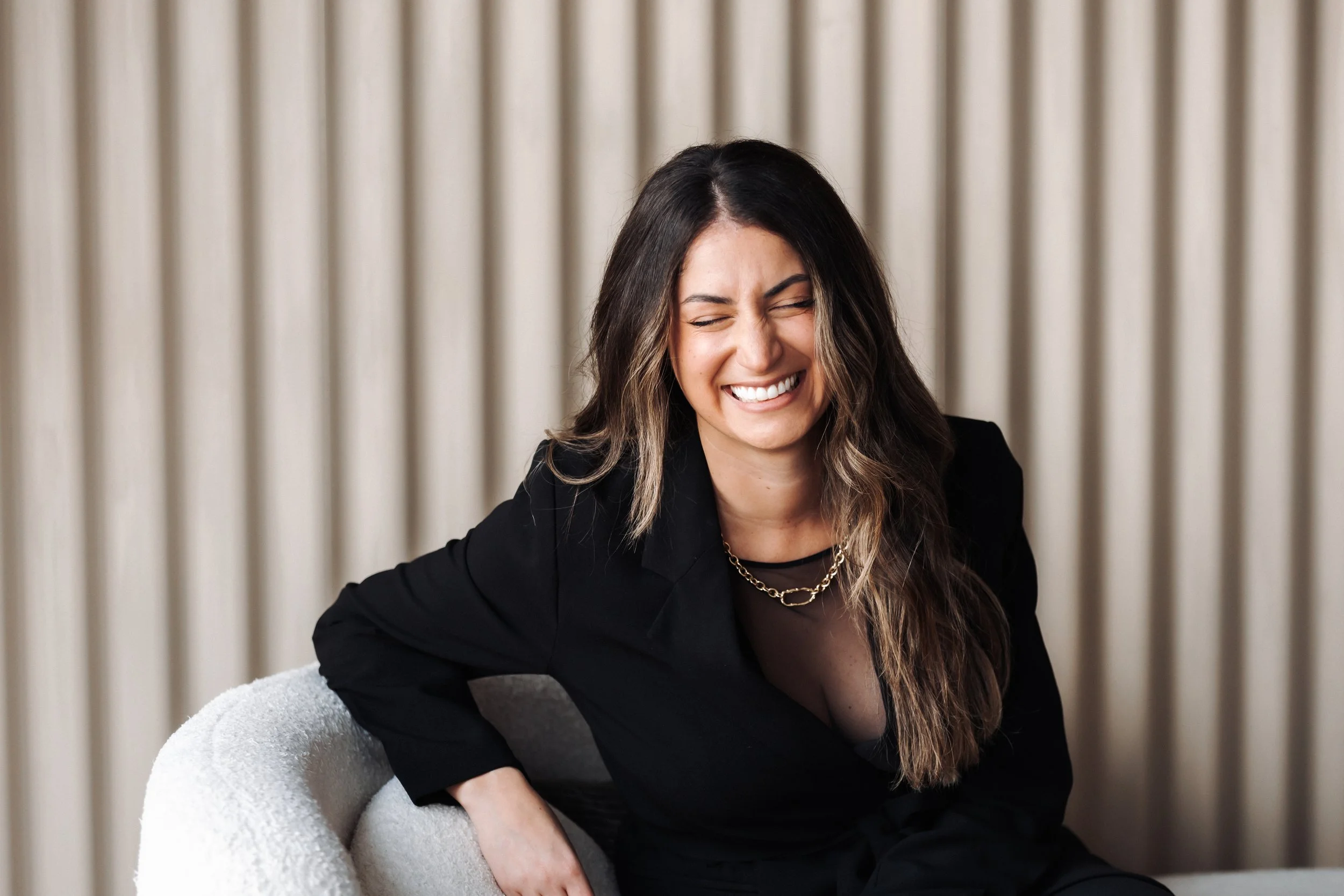 A woman with long brown hair and light skin, wearing a black blazer and gold chain necklace, smiling with her eyes closed while sitting on a light-colored armchair against a beige curtain background.