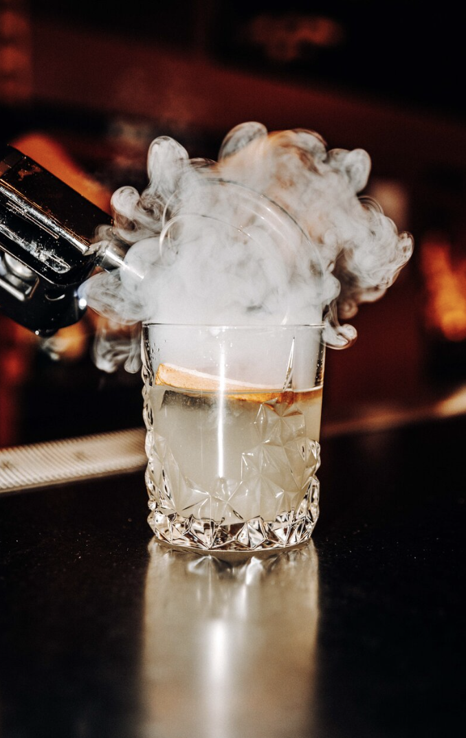 A glass with a drink and a lemon slice, with dry ice creating fog overflowing from the glass, on a dark bar counter.