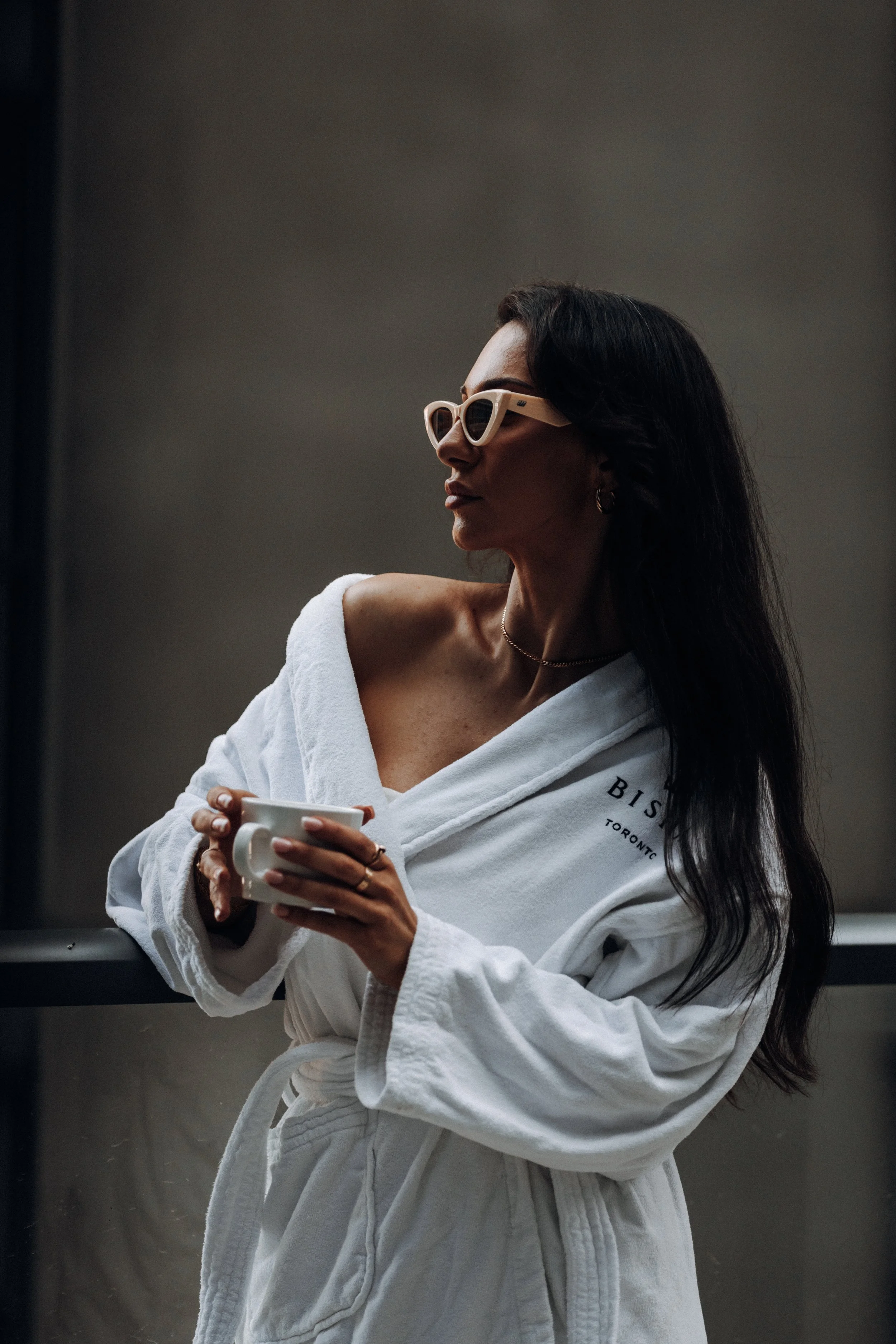 Woman with dark hair wearing glasses and a white robe, holding a white mug, standing in a dimly lit space.